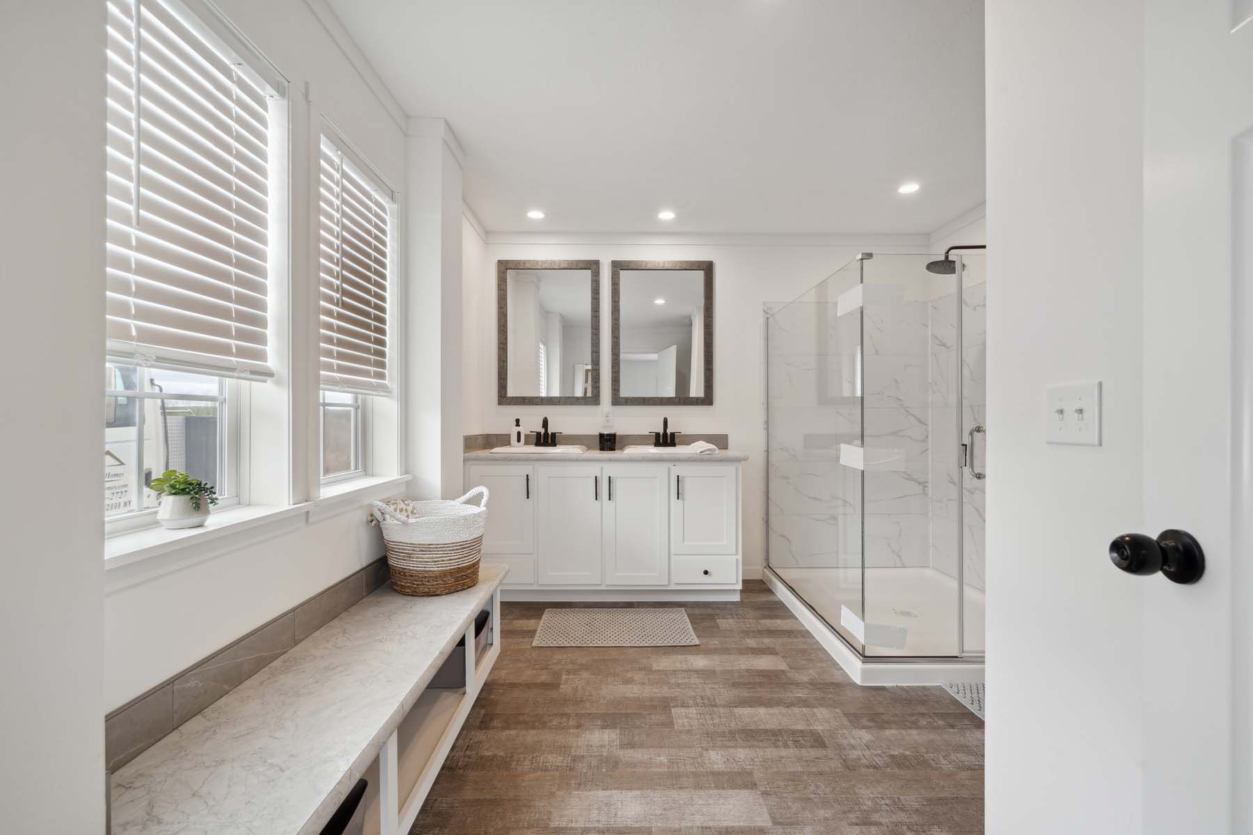 Bright, modern bathroom with large windows, wooden flooring, and a glass shower. Double sinks under two mirrors, a bench with storage, and a basket.