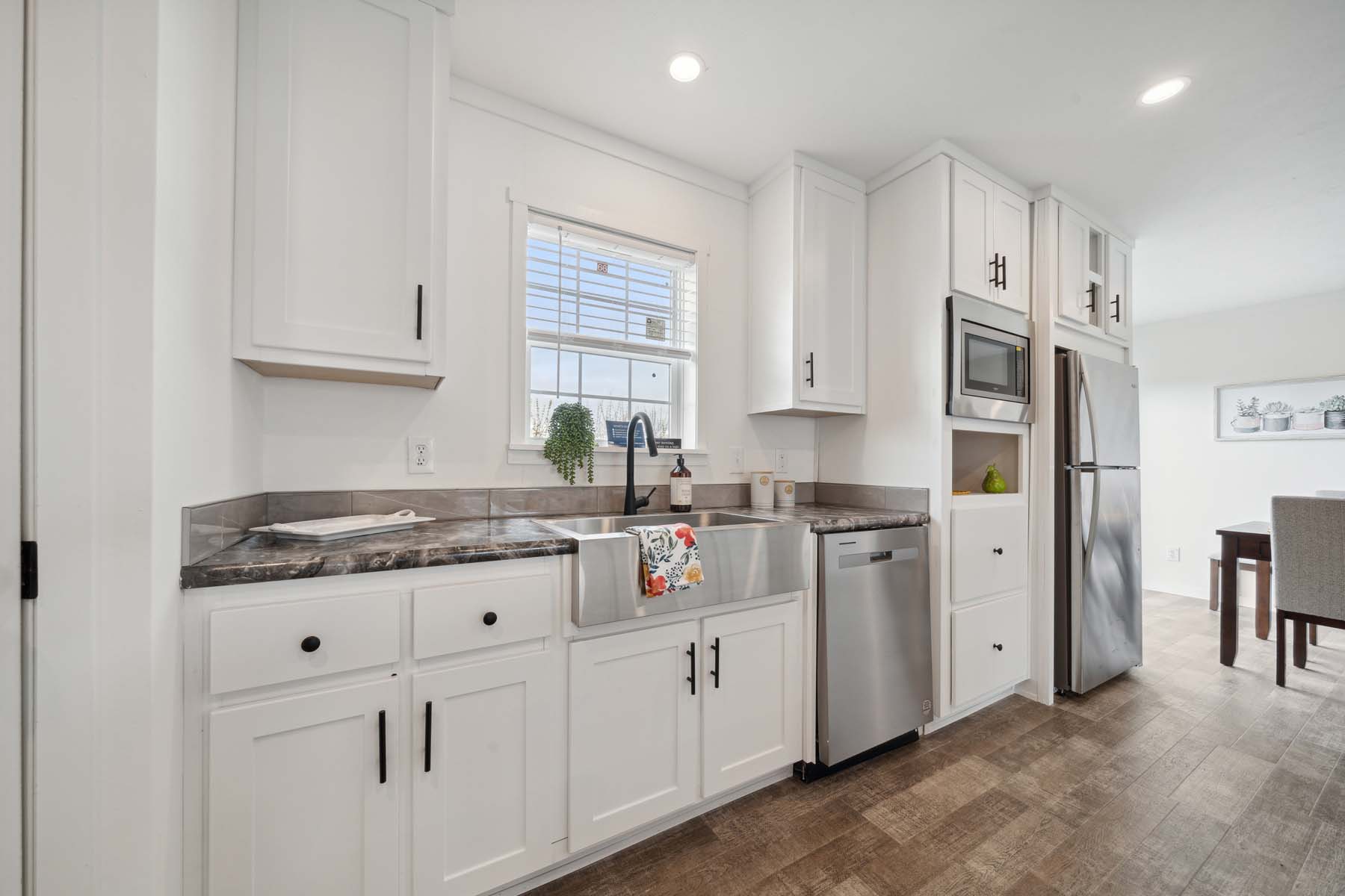 Bright kitchen with white cabinets, a farmhouse sink, and stainless steel appliances. A window with a potted plant allows natural light, creating a fresh feel.