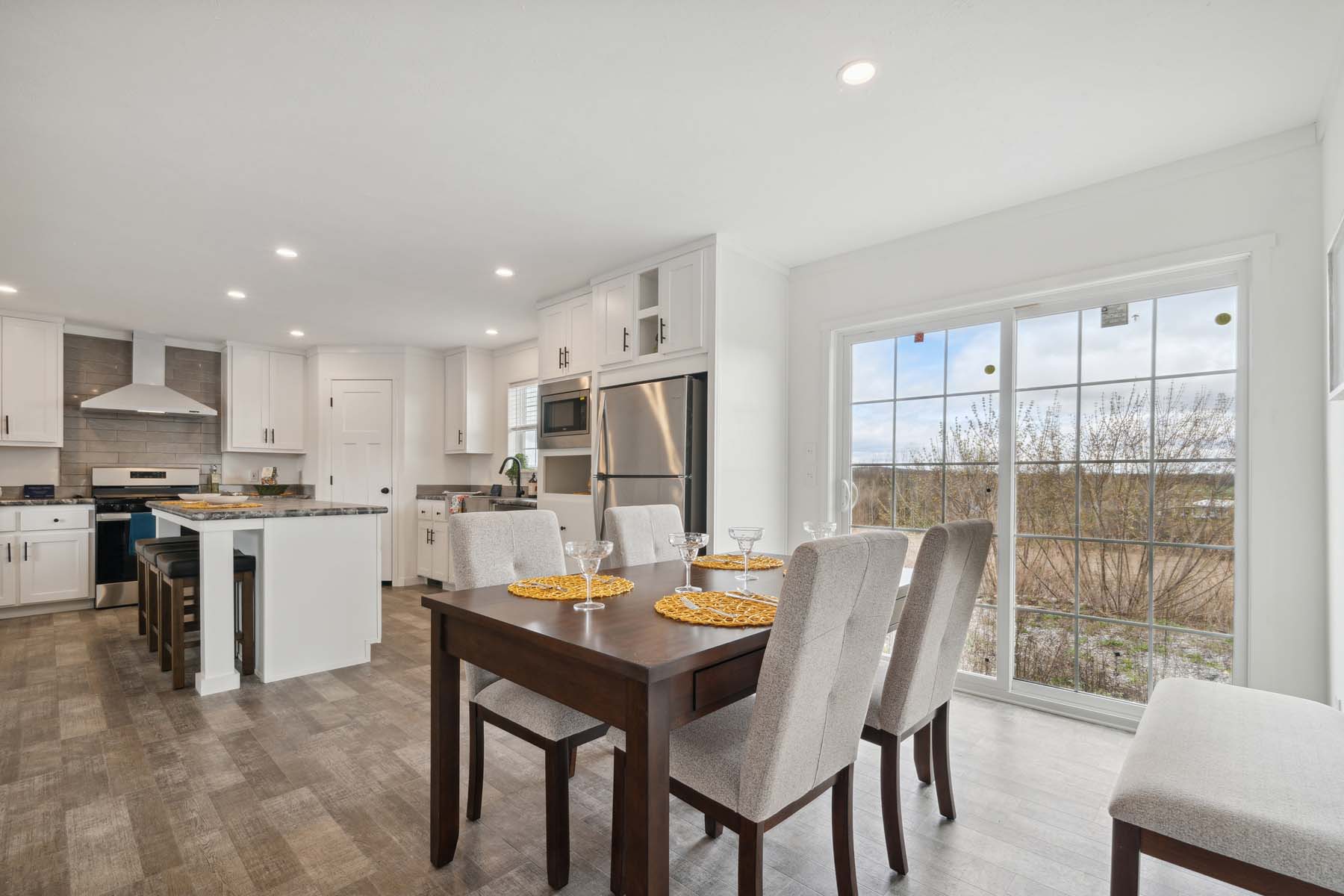 Modern kitchen and dining area with a wooden table set for four, gray chairs, and a window view. The kitchen features stainless steel appliances.