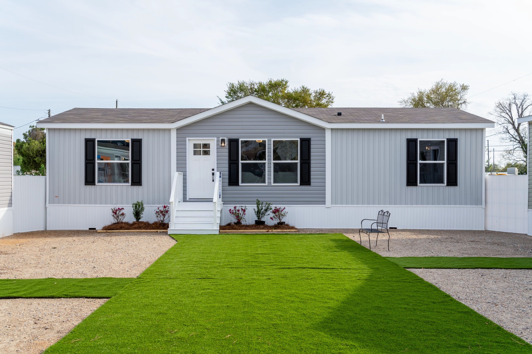 A modern gray and white manufactured home with a gabled roof, black shutters, and a small front porch. Neat, artificial green grass and gravel pathways create a tidy yard.