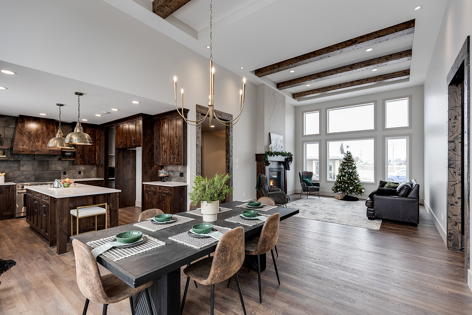 Modern open-concept living area with a brown kitchen, cozy dining table set for four, and a living room featuring a decorated Christmas tree by large windows.