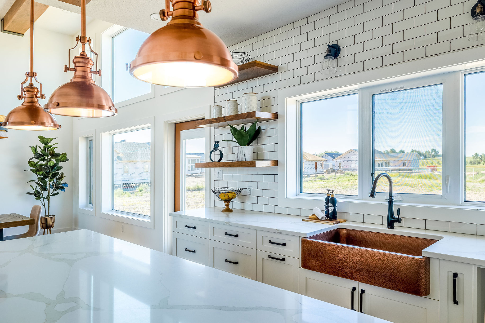 Modern kitchen with natural light, featuring copper pendant lights, white cabinetry, a copper sink, and green plants on wooden shelves. Bright and airy.