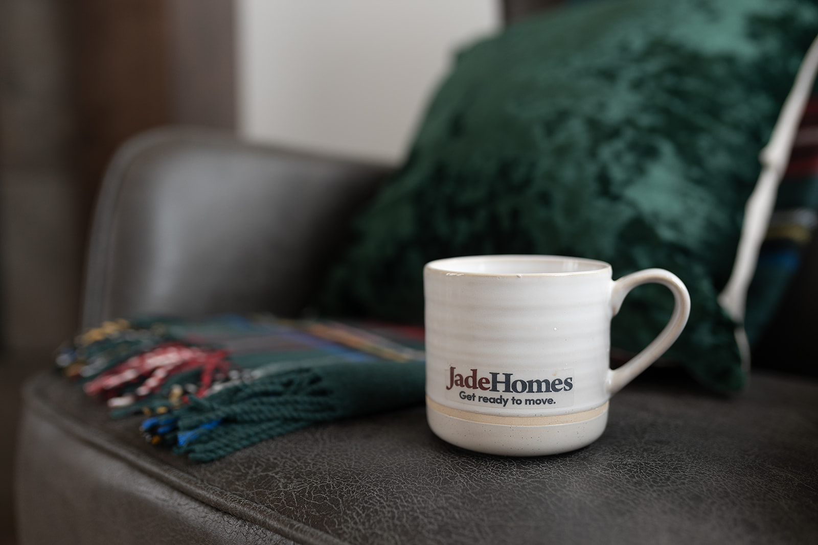 A white mug with "Jade Homes" text sits on a gray sofa. Behind it is a green pillow and a fringed, colorful blanket, creating a cozy atmosphere.