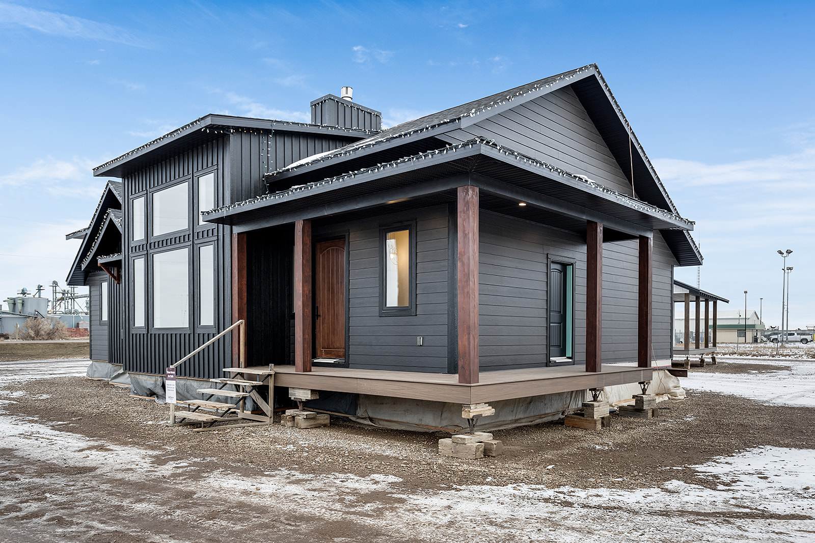 Modern prefab house with dark siding, large windows, and a wooden porch on a snowy, muddy lot. The sky is clear blue, creating a crisp, serene atmosphere.