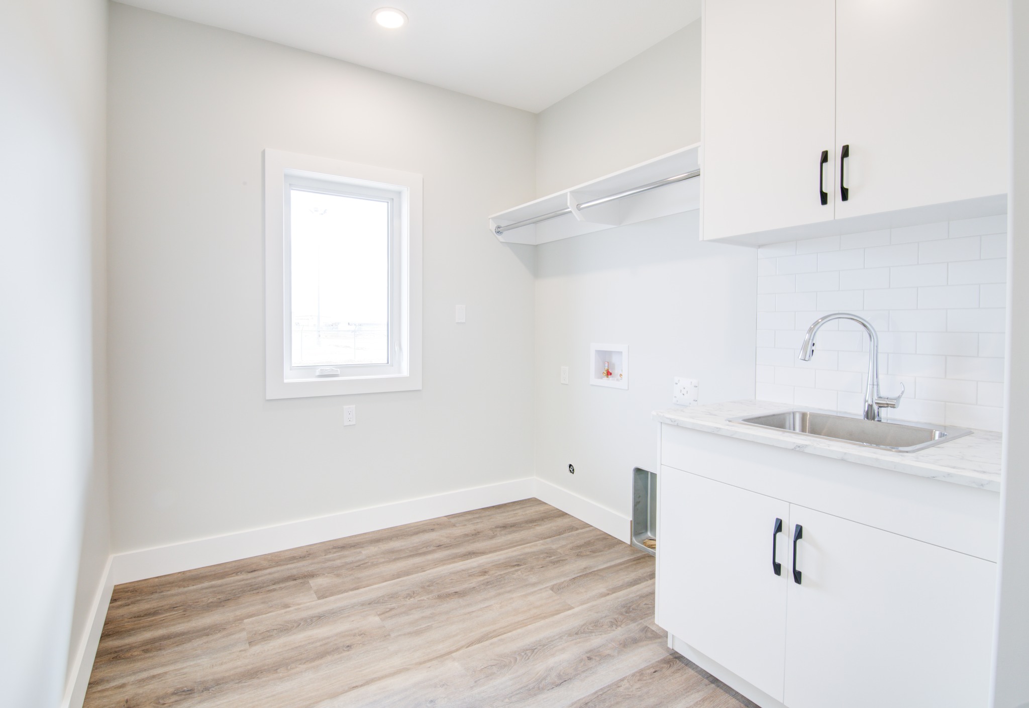 Bright laundry room with a single window, light wood floors, white cabinetry, subway tile backsplash, and a stainless steel sink, conveying a clean, modern feel.