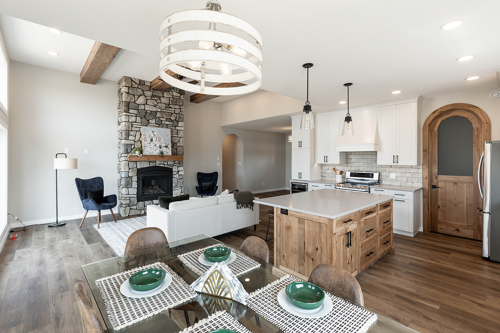 Spacious, modern open-plan kitchen and living room. Features stone fireplace, wooden beams, sleek white cabinets, and a glass dining table. Cozy and inviting.