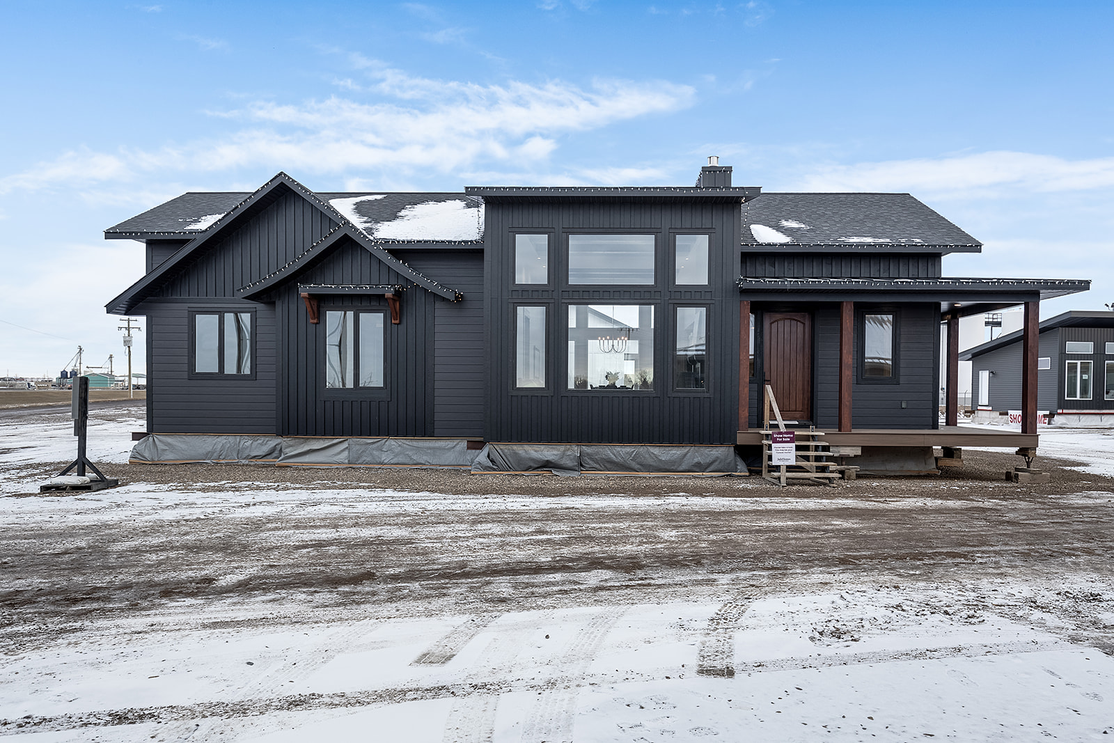 Modern dark gray house with large windows, set on a snowy landscape under a clear blue sky. The building appears new and unoccupied.