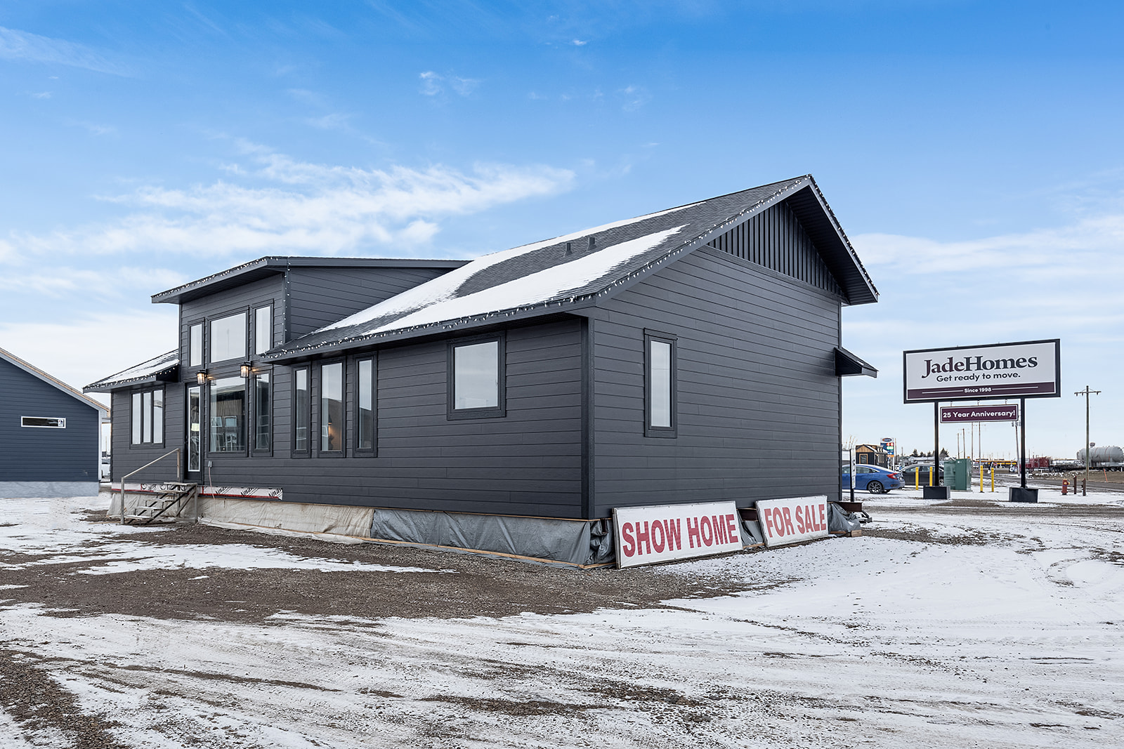 Gray show home in snowy landscape with large windows and a sloped roof. Signs read "Show Home For Sale." A commercial sign is visible in the background.