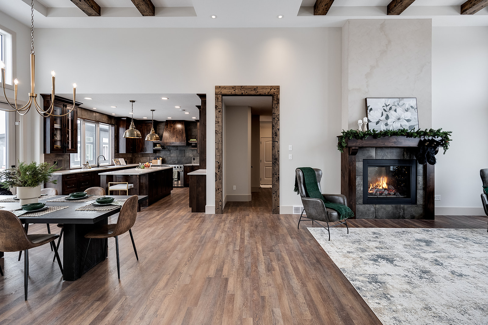 Spacious, modern living-dining area with a warm fireplace on the right, sleek kitchen on the left, and a rustic dining table set in the foreground. Cozy and elegant.