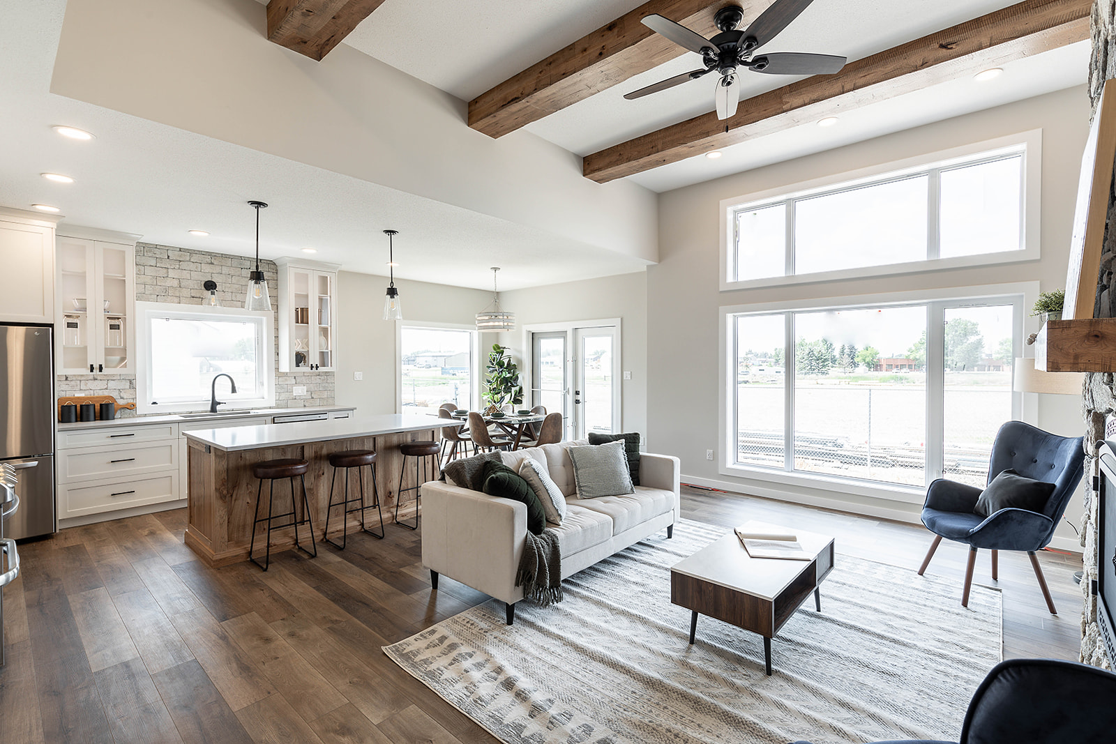 Modern open-concept living area with wooden beams, cozy sofa, and kitchen island. Large windows provide natural light, creating an airy, inviting atmosphere.