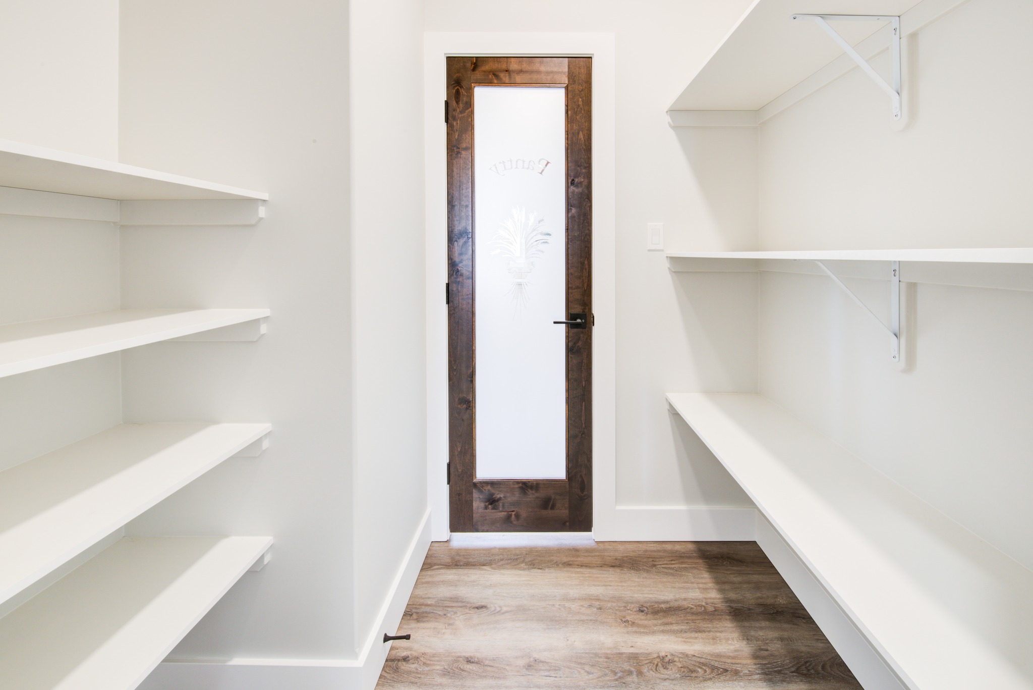 White pantry with wooden flooring, featuring empty white shelves on both sides. A wooden-framed glass door at the end adds a warm touch.