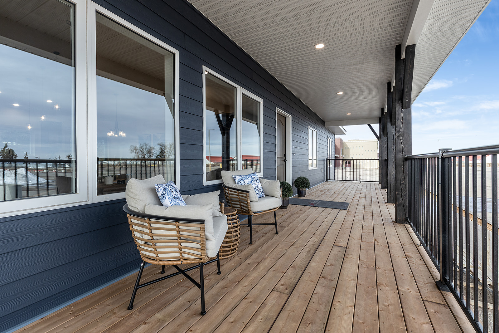 Spacious wooden porch with two wicker chairs, cream cushions, and blue pillows against a dark blue wall and large windows. Bright, relaxing atmosphere.