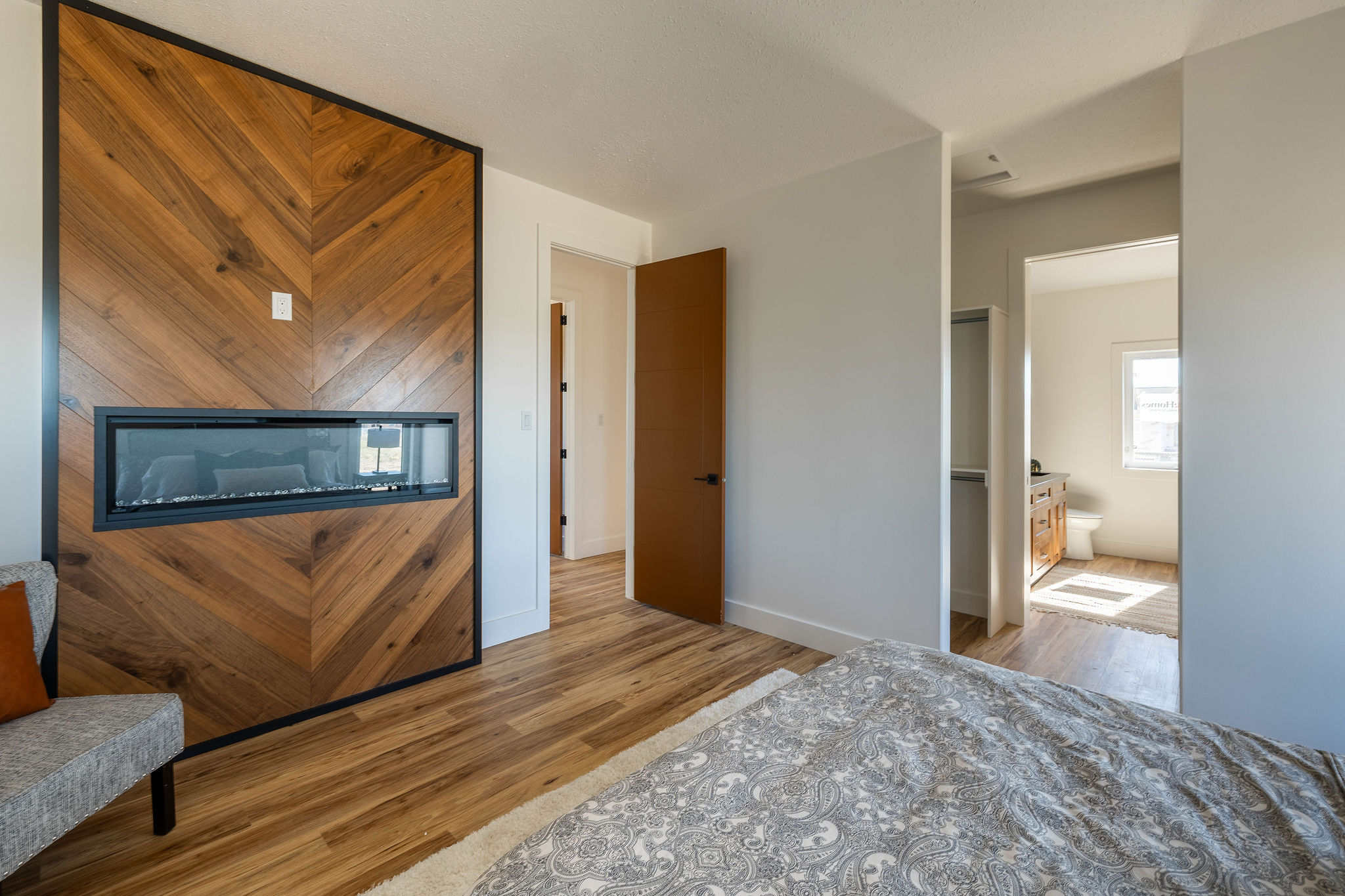 Modern bedroom with a chevron wood accent wall and electric fireplace. Open door reveals light-filled bathroom. Neutral tones create a cozy ambiance.