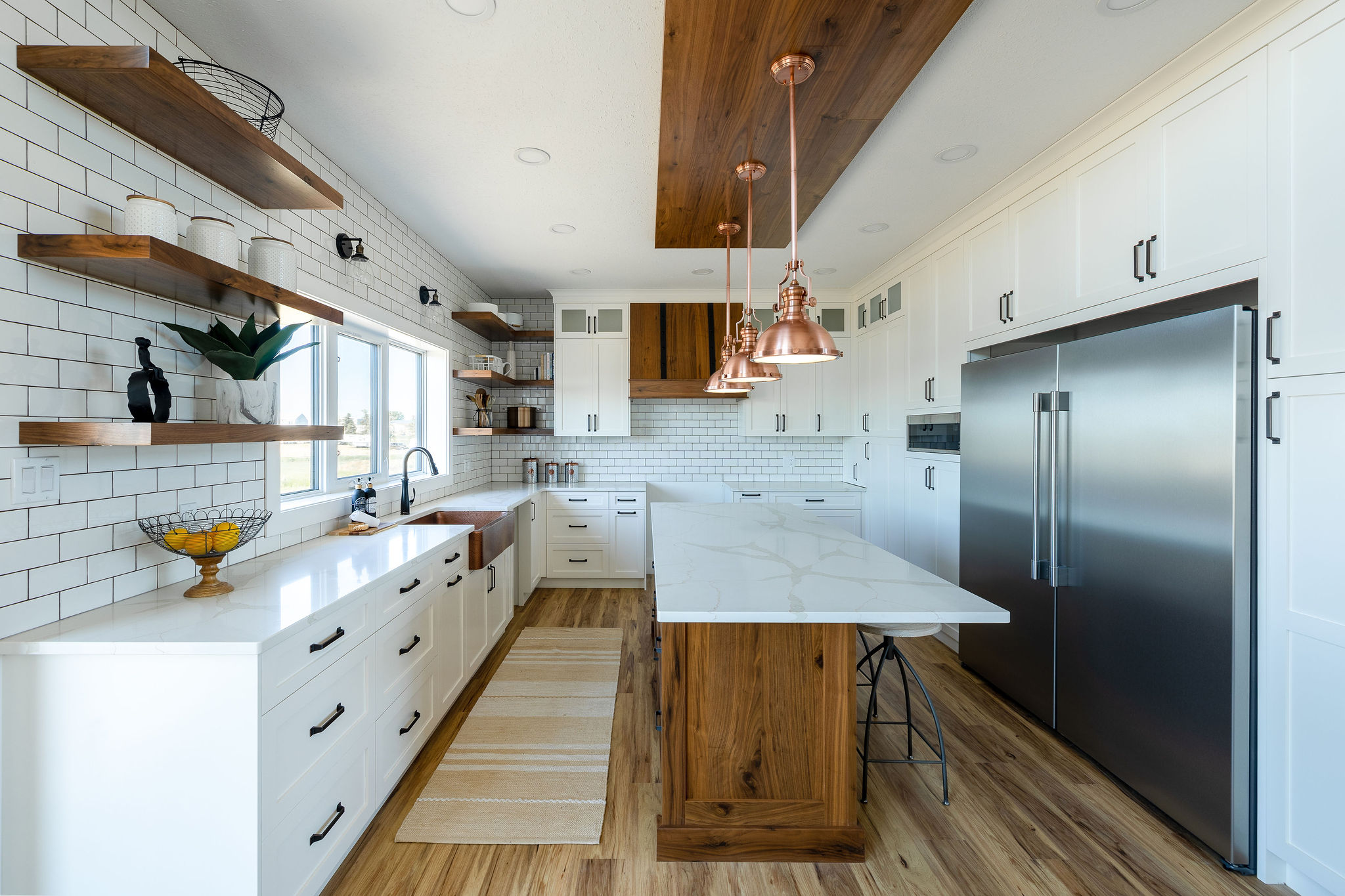Modern kitchen with white cabinets, wood accents, and copper pendant lights. Open shelves hold decor items. A large stainless steel fridge is on the right.