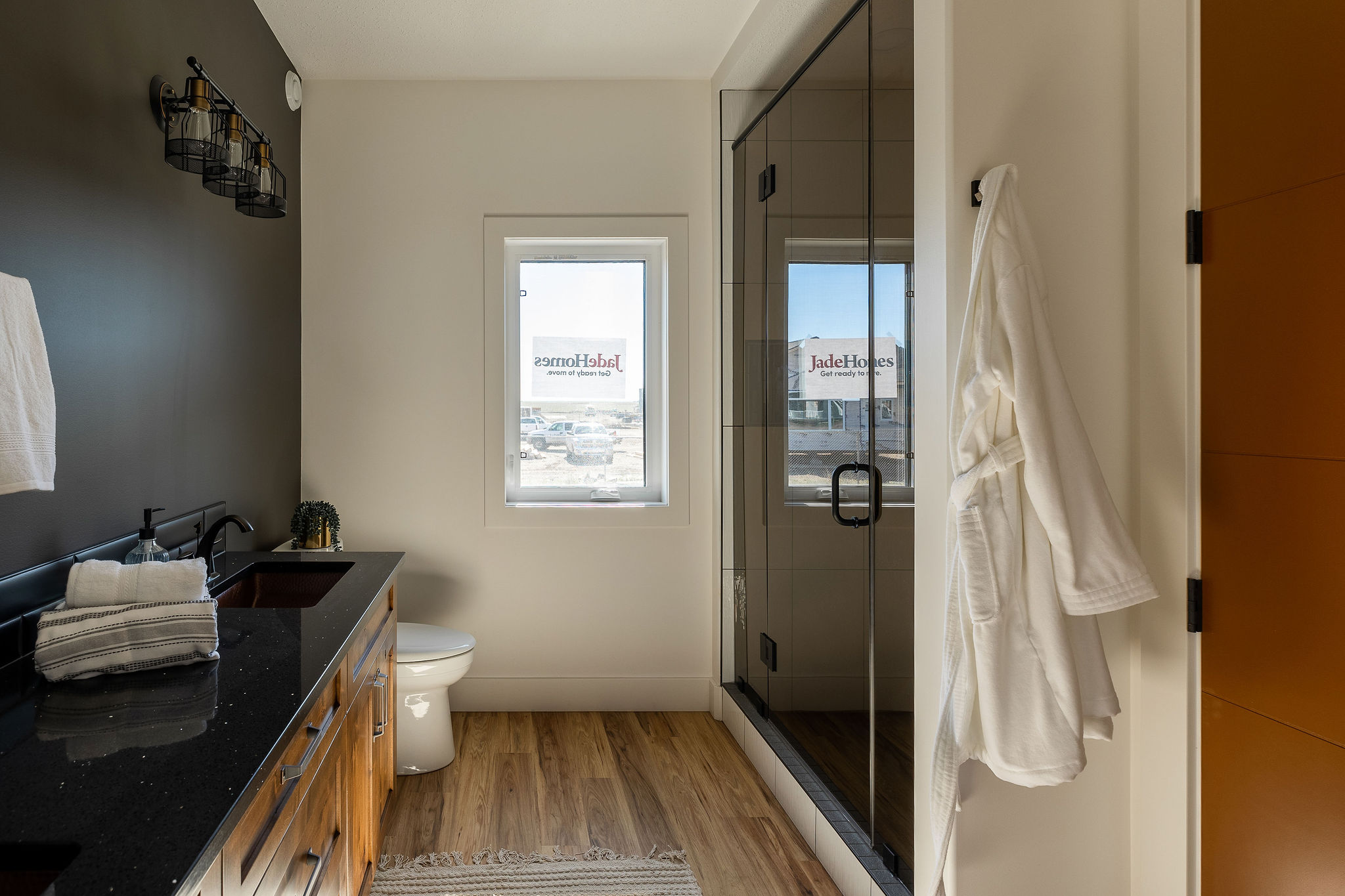 Modern bathroom with dark countertop, wood cabinets, and a black faucet. Glass shower door, window with view, and white robe on the door. Calm, elegant feel.