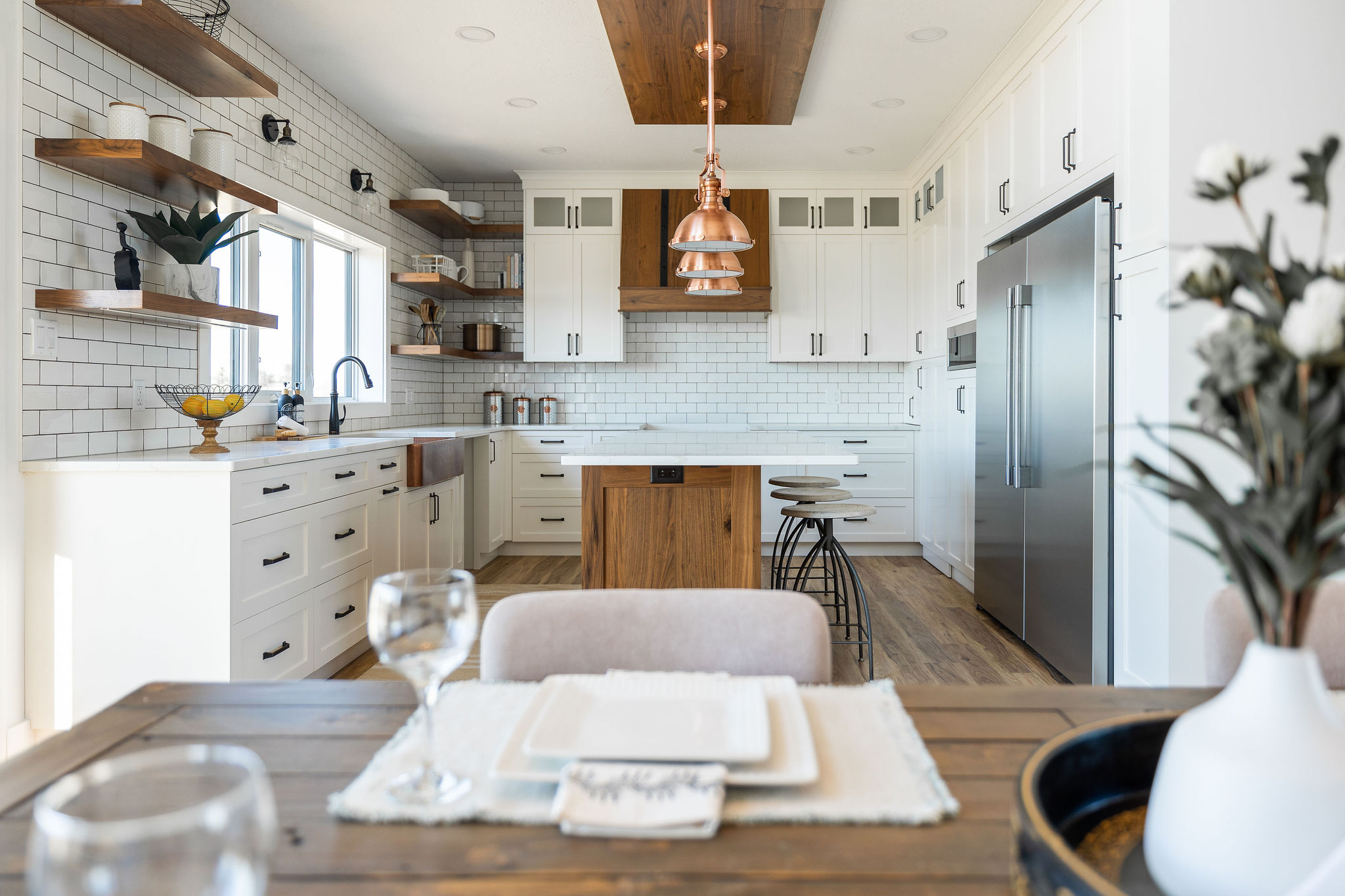 Bright, modern kitchen with white cabinets, wooden island, and copper pendant lights. Open shelving, subway tiles, and elegant dining table in foreground.