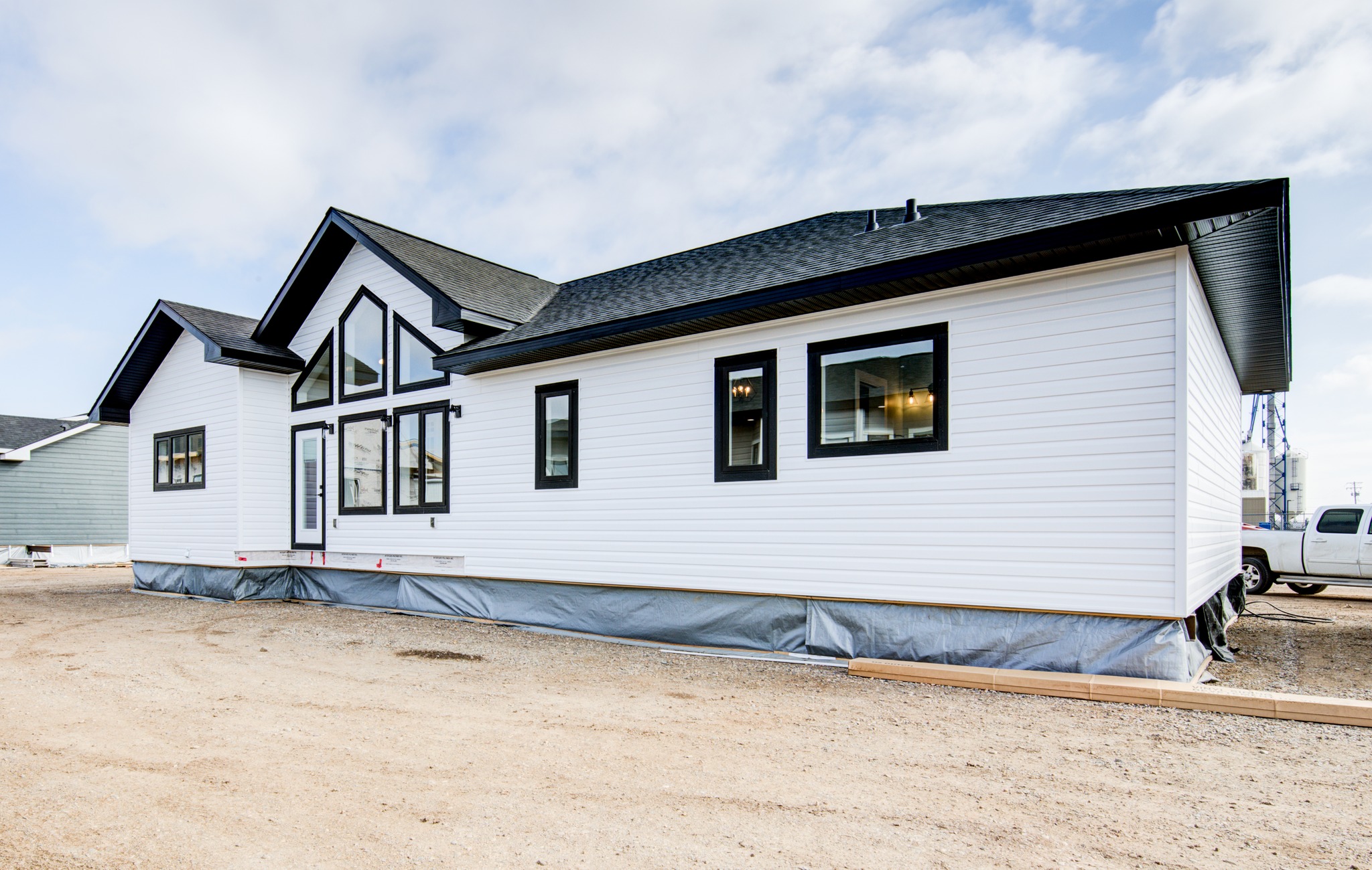 Modern white modular home with black trim, large windows, and a sloped roof on a gravel lot. Overcast sky, soft light, and a truck parked nearby.