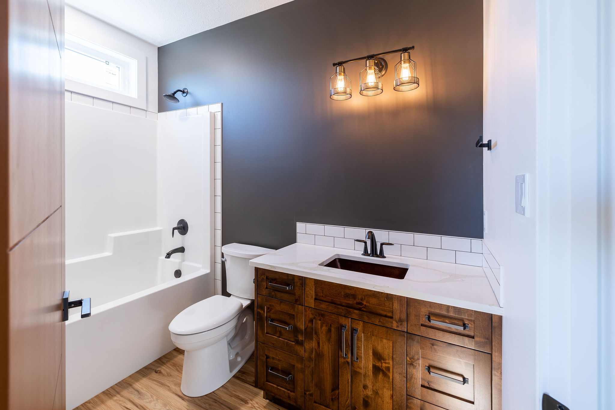 Modern bathroom with gray walls and warm lighting. Features a wooden vanity with white countertop, a white bathtub, toilet, and wood floor. Cozy ambiance.