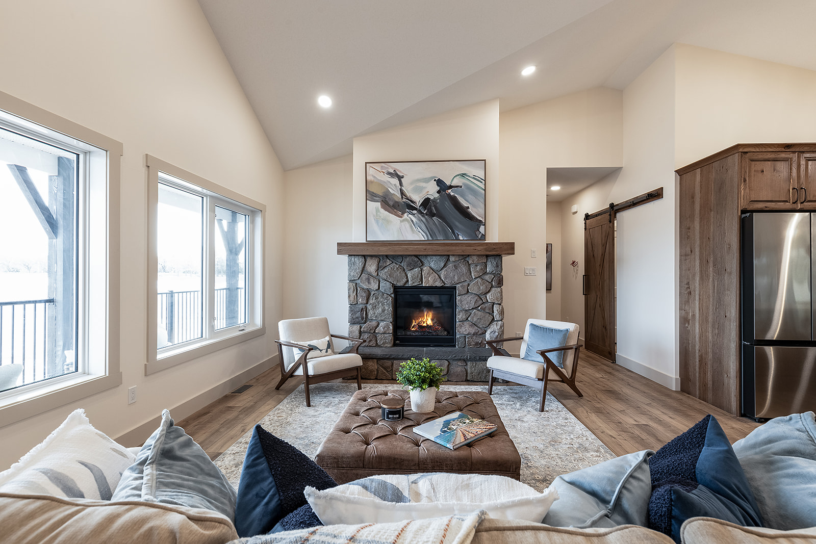 Cozy living room with a stone fireplace, modern art above. Soft chairs flank the fire; a plush ottoman, magazines, and plant add warmth and comfort.