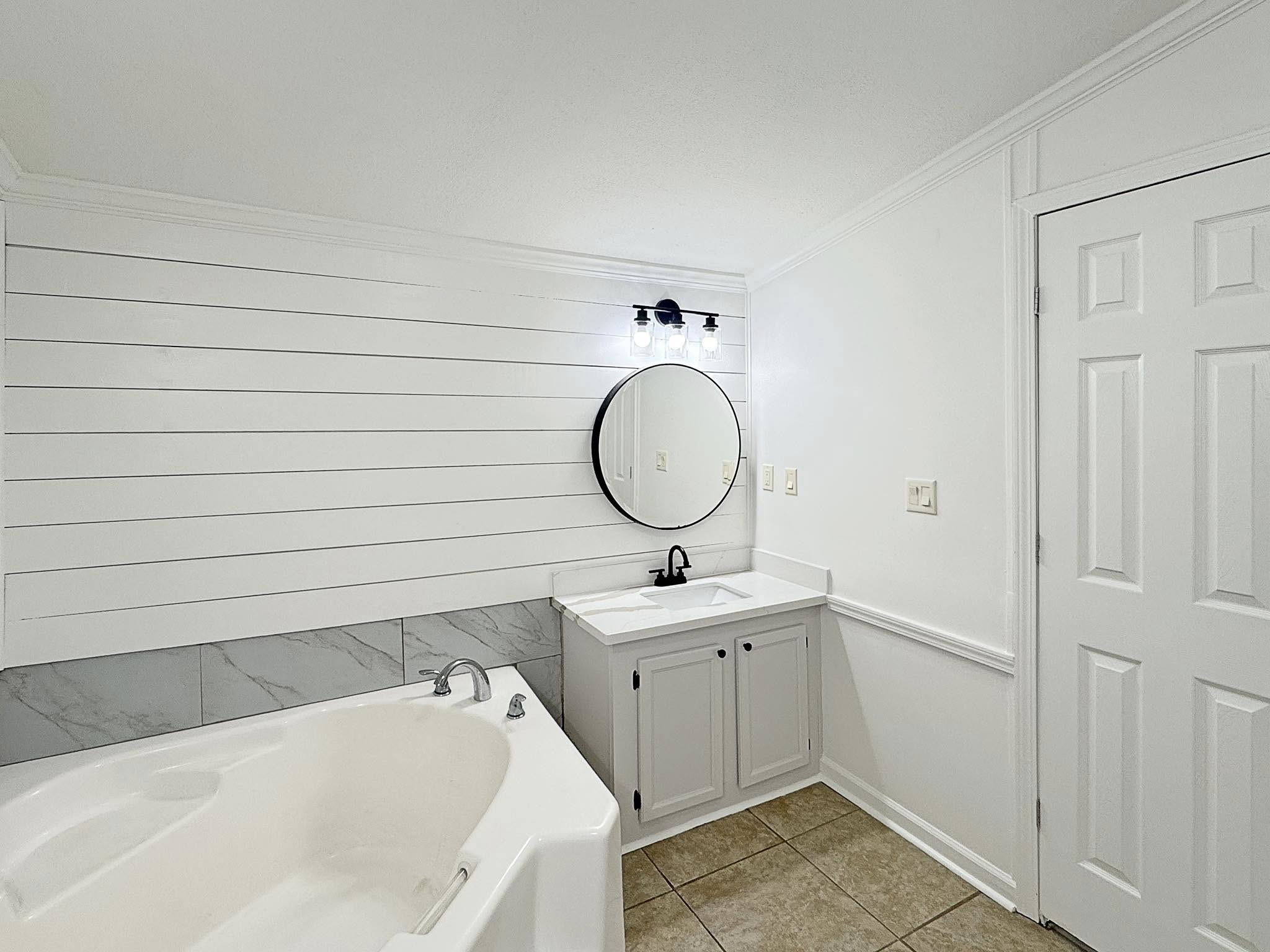 Bright bathroom with white shiplap walls features a corner bathtub and a vanity. A round mirror and black light fixture add modern touches.