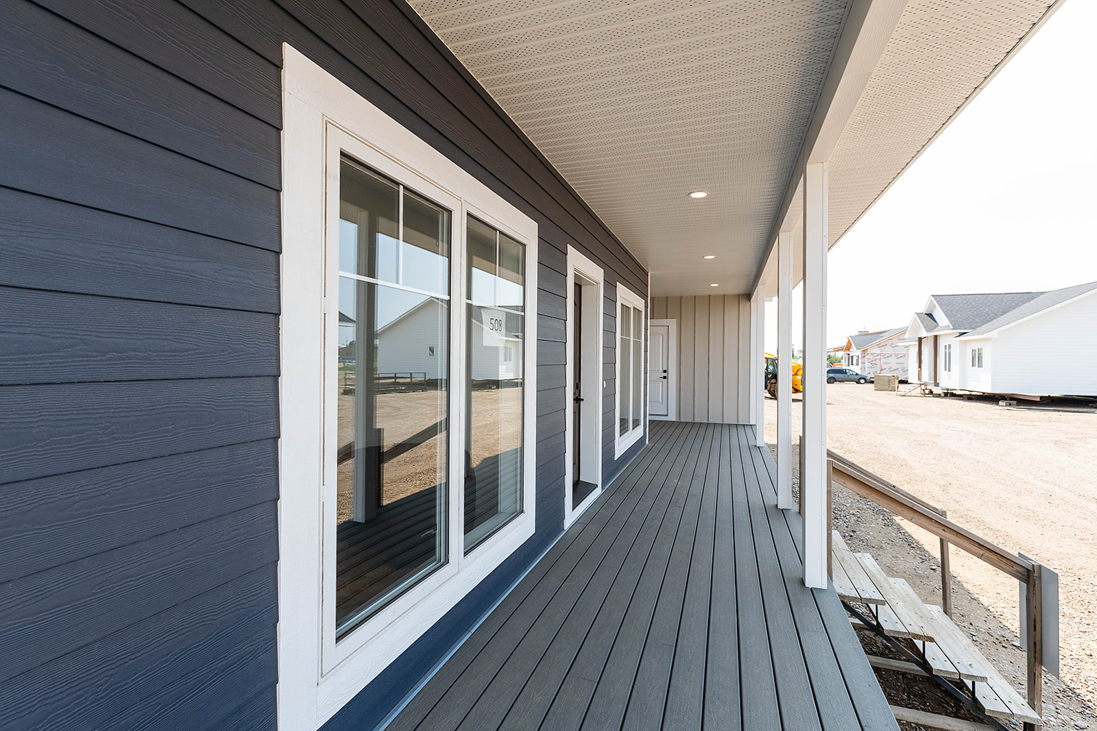 Wide porch with gray decking, white-trimmed windows, and blue siding. Overhead lights highlight a clean, industrial look; nearby homes visible.
