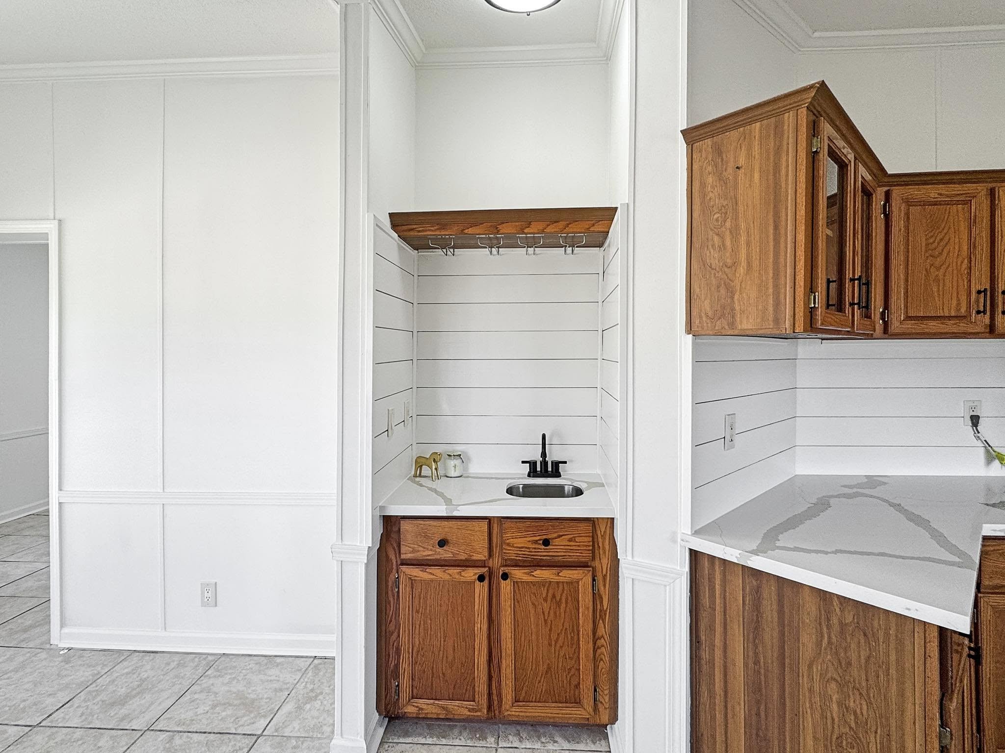 A compact kitchenette with a small sink, brass faucet, and white shiplap walls. Wood cabinets and a marble countertop add warmth to the minimalist space.