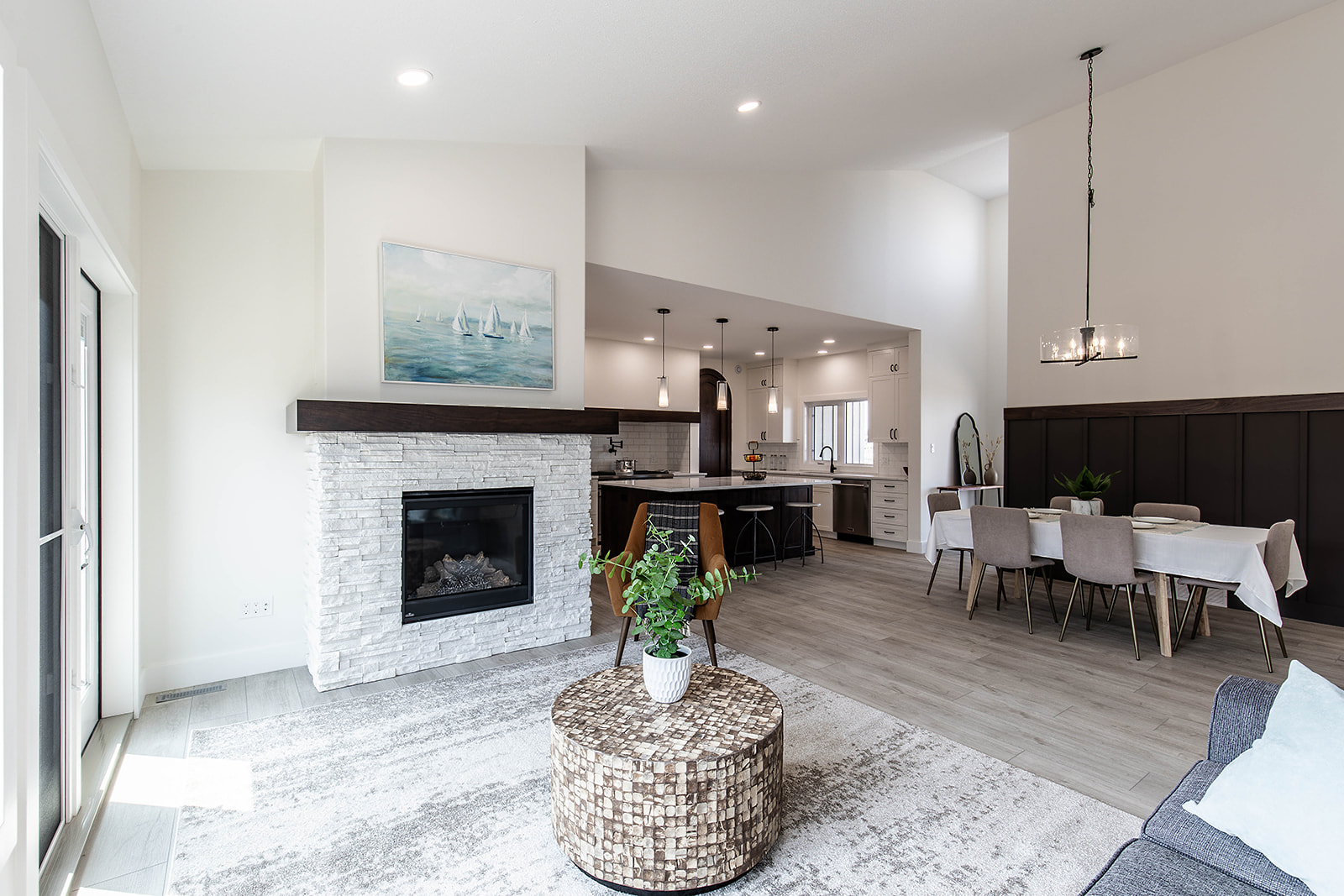 Modern living room with high ceilings, light gray walls, and wood flooring. Features a stone fireplace, round coffee table, and cozy dining area.