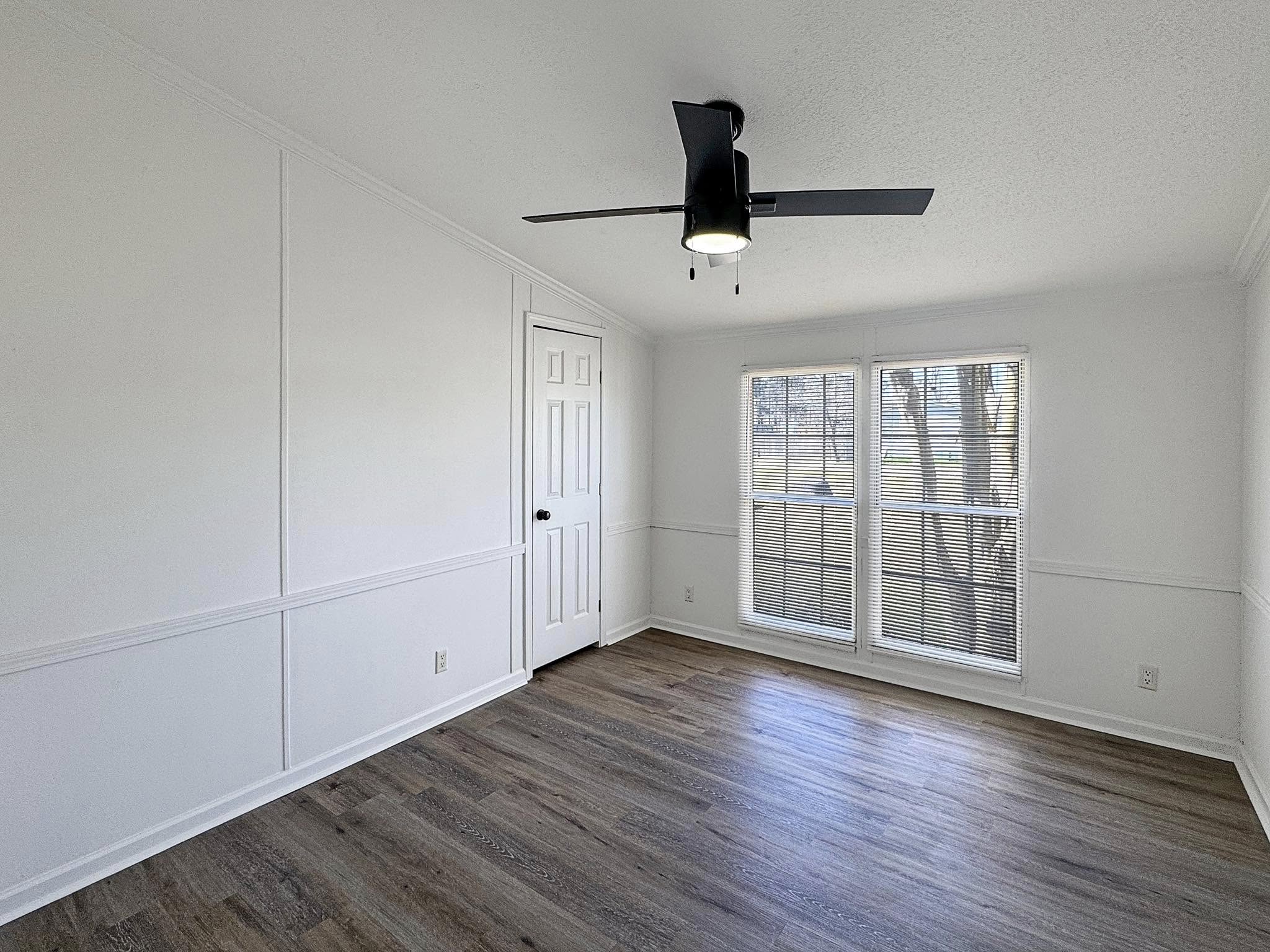 Empty room with white walls, a black ceiling fan, and wood flooring. Sunlight filters through two tall windows with blinds, creating a serene atmosphere.