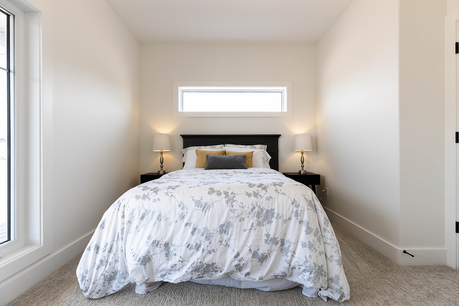 A cozy bedroom features a bed with a white and gray floral comforter, flanked by two lamps on dark nightstands. Soft natural light streams through a window.