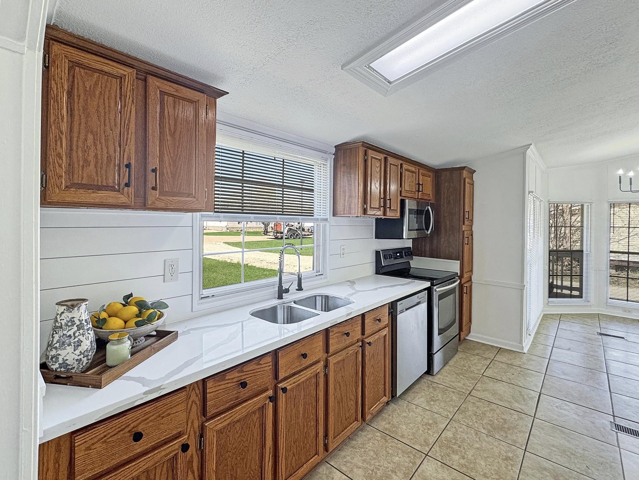 Bright kitchen with wooden cabinets, white countertops, and stainless steel appliances. A window above the sink reveals a sunny view. Fresh fruit and a vase decorate the counter, creating a cozy, inviting atmosphere.