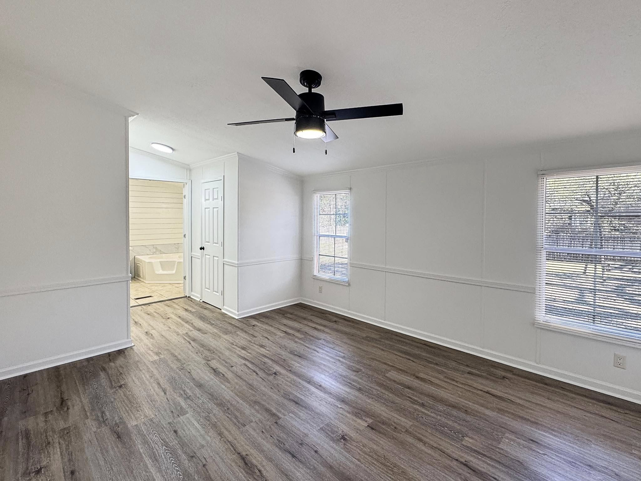 Spacious, bright room with white walls and wood flooring. A ceiling fan hangs overhead. Two large windows on the right let in natural light. A bathroom is visible through a doorway in the back.