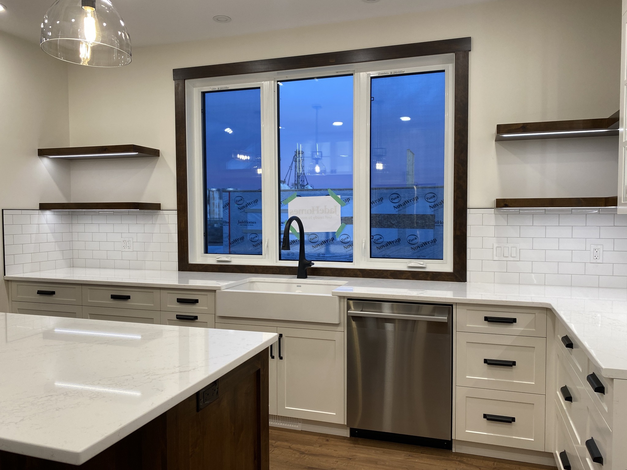 Modern kitchen with white cabinets and black handles, featuring a large window framed in dark wood. There’s a farmhouse sink, stainless steel dishwasher, and open shelves. Bright, clean ambiance with a warm wood floor and sleek countertops.