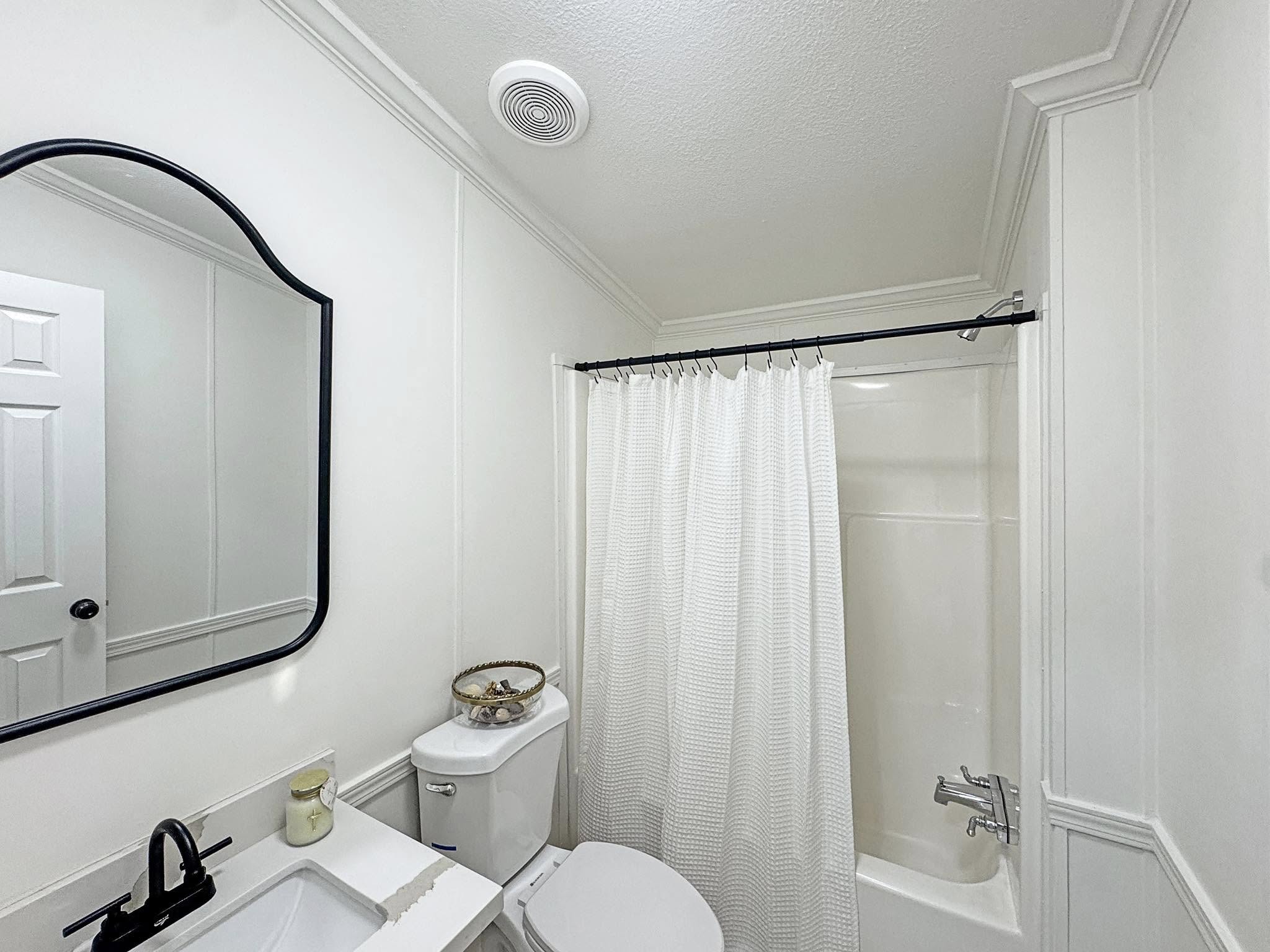 A small, clean bathroom with a white bathtub and shower curtain, white toilet, black faucet, rectangular sink, and a wall mirror. Minimalist decor.