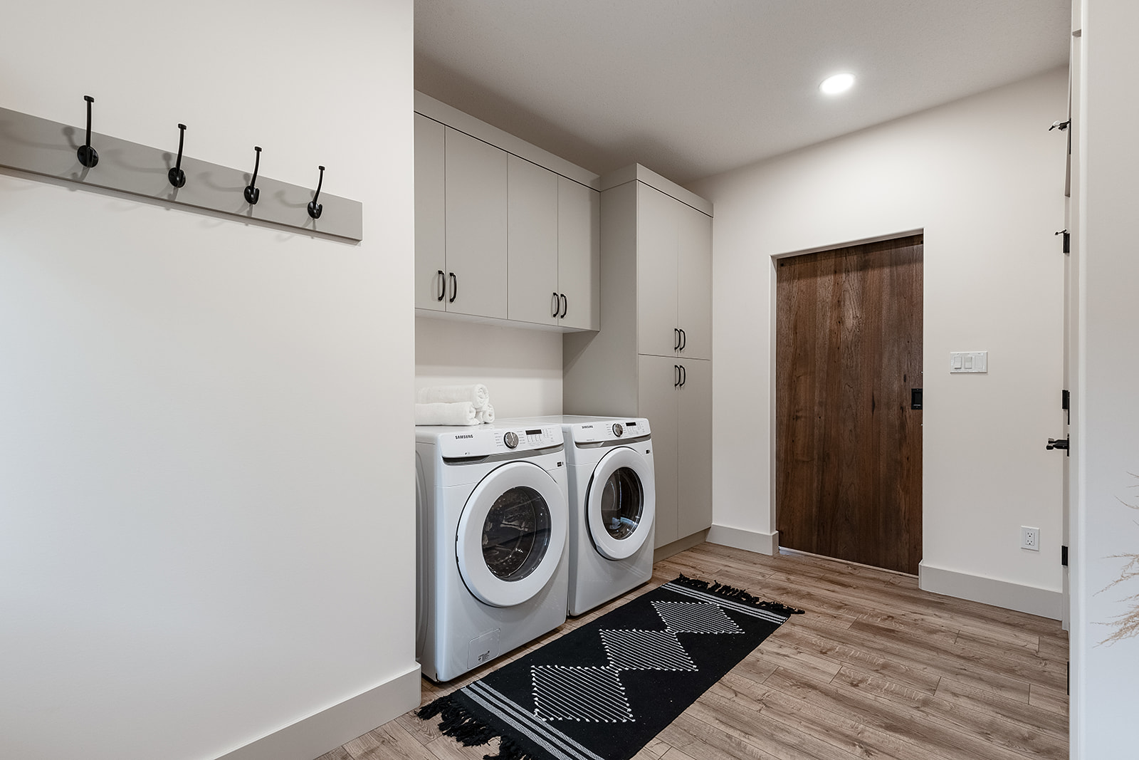 Modern laundry room with stacked washer and dryer, light wood floor, dark rug, white cabinets, and a wooden sliding door, conveying cleanliness.