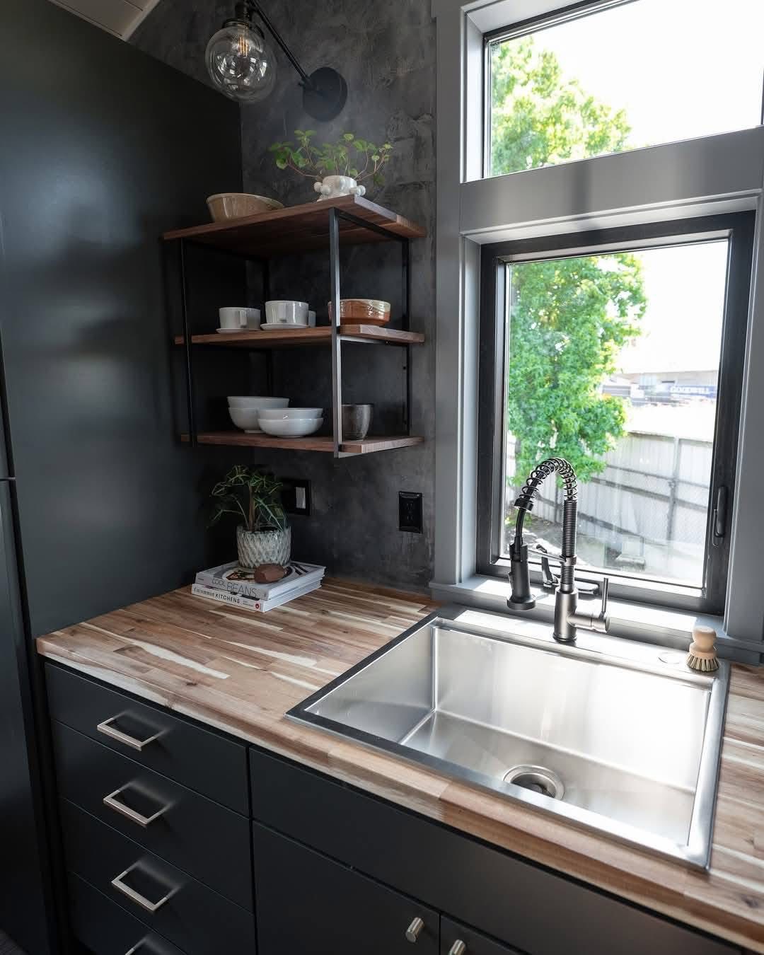 Modern kitchen corner with sleek, dark cabinets, a wooden countertop, and a metal sink. Open shelves hold dishes; a window offers a garden view. Cozy and organized.