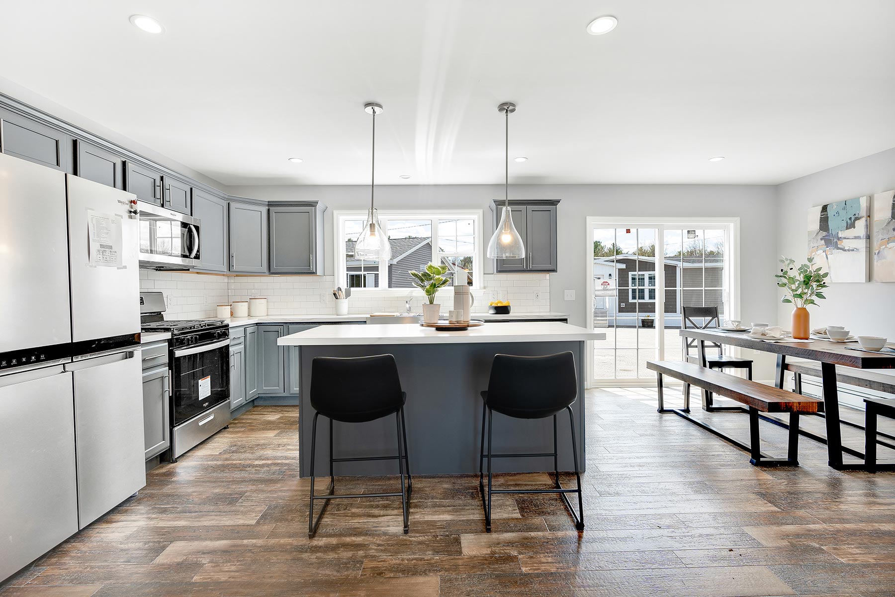 Modern kitchen with gray cabinets, white countertops, and wood floors. A center island has two black chairs and pendant lights. Dining area with bench seating. Bright and airy atmosphere.
