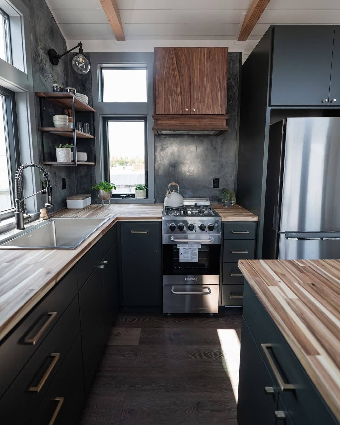 Modern kitchen with dark cabinets and wood countertops. Features a stainless steel oven, fridge, and a large sink. Open shelves hold plants and dishes.