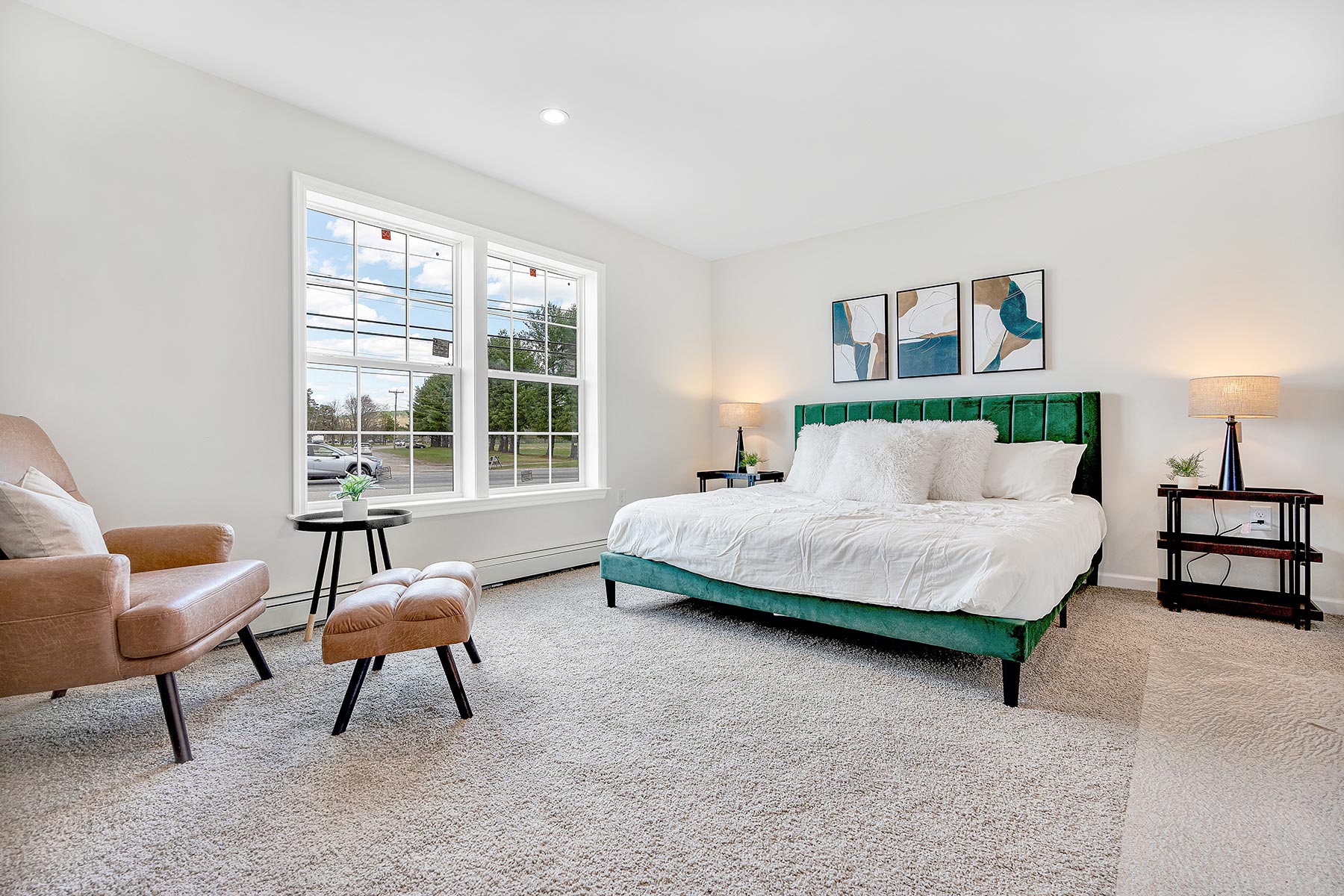 Bright bedroom with a green velvet bed, white bedding, and abstract wall art. A cozy tan armchair and footstool sit near a large window. Airy, calm atmosphere.