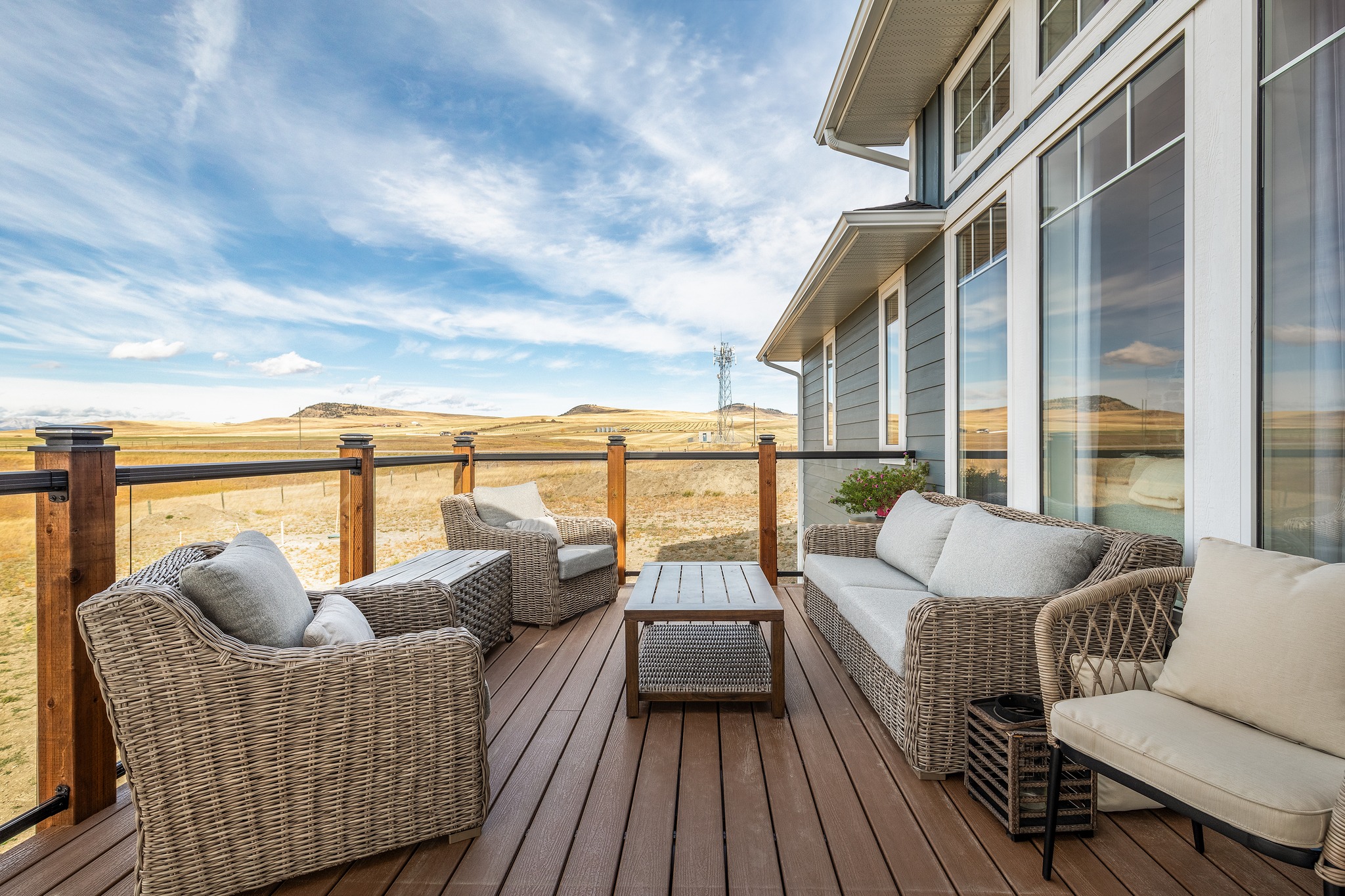 Exterior view of a home deck with wicker furniture, cream cushions, and a wooden railing. The setting overlooks a serene, open landscape under a blue sky.