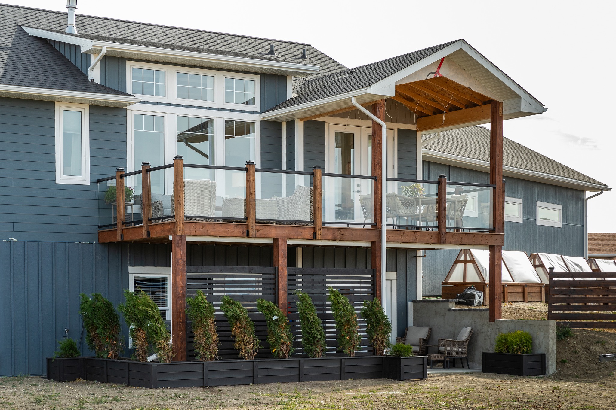 Two-story blue house with a wooden deck and glass railing. Below, a row of neatly arranged potted plants adds greenery. Cozy patio furniture is visible.