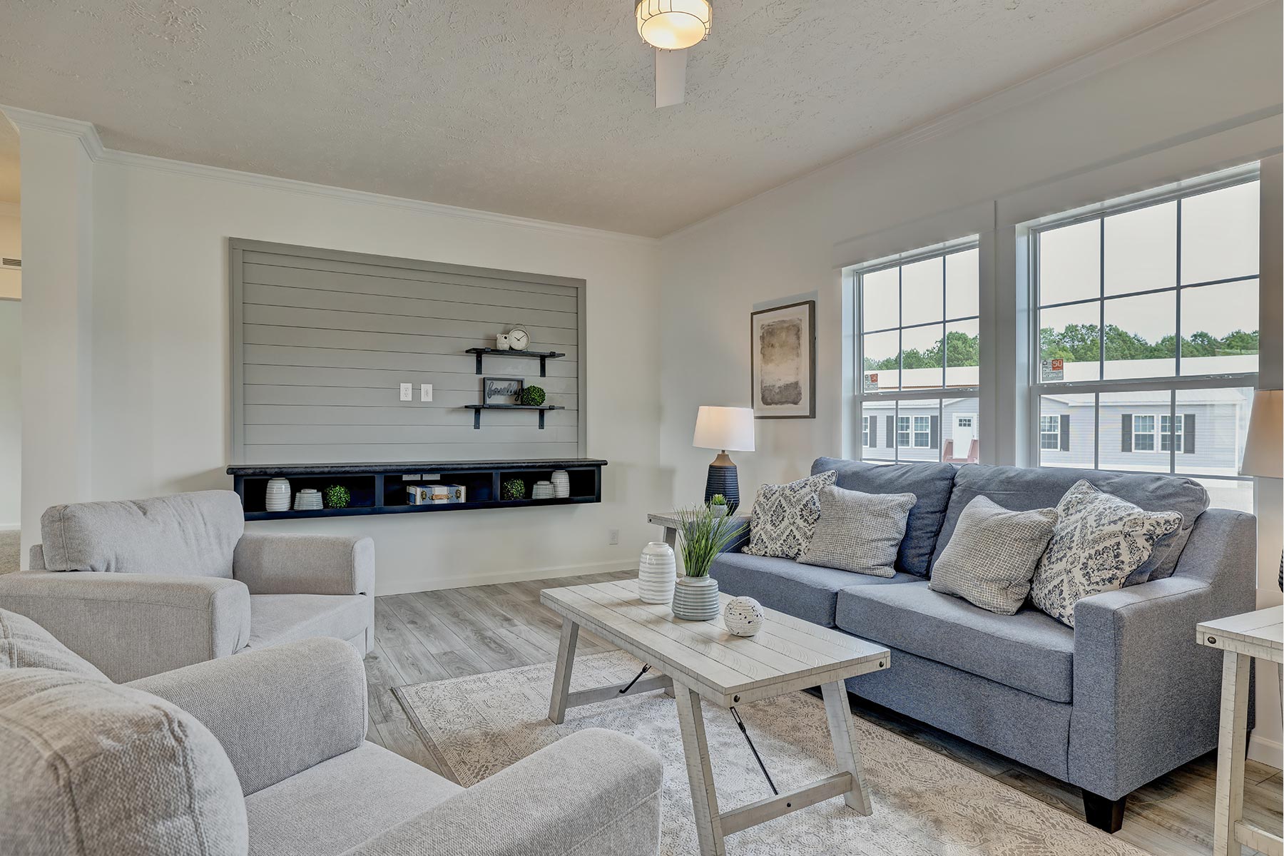 Cozy living room with a gray sofa and matching armchairs, decorative pillows, and a wooden coffee table. Large windows and shelves add a bright, inviting touch.