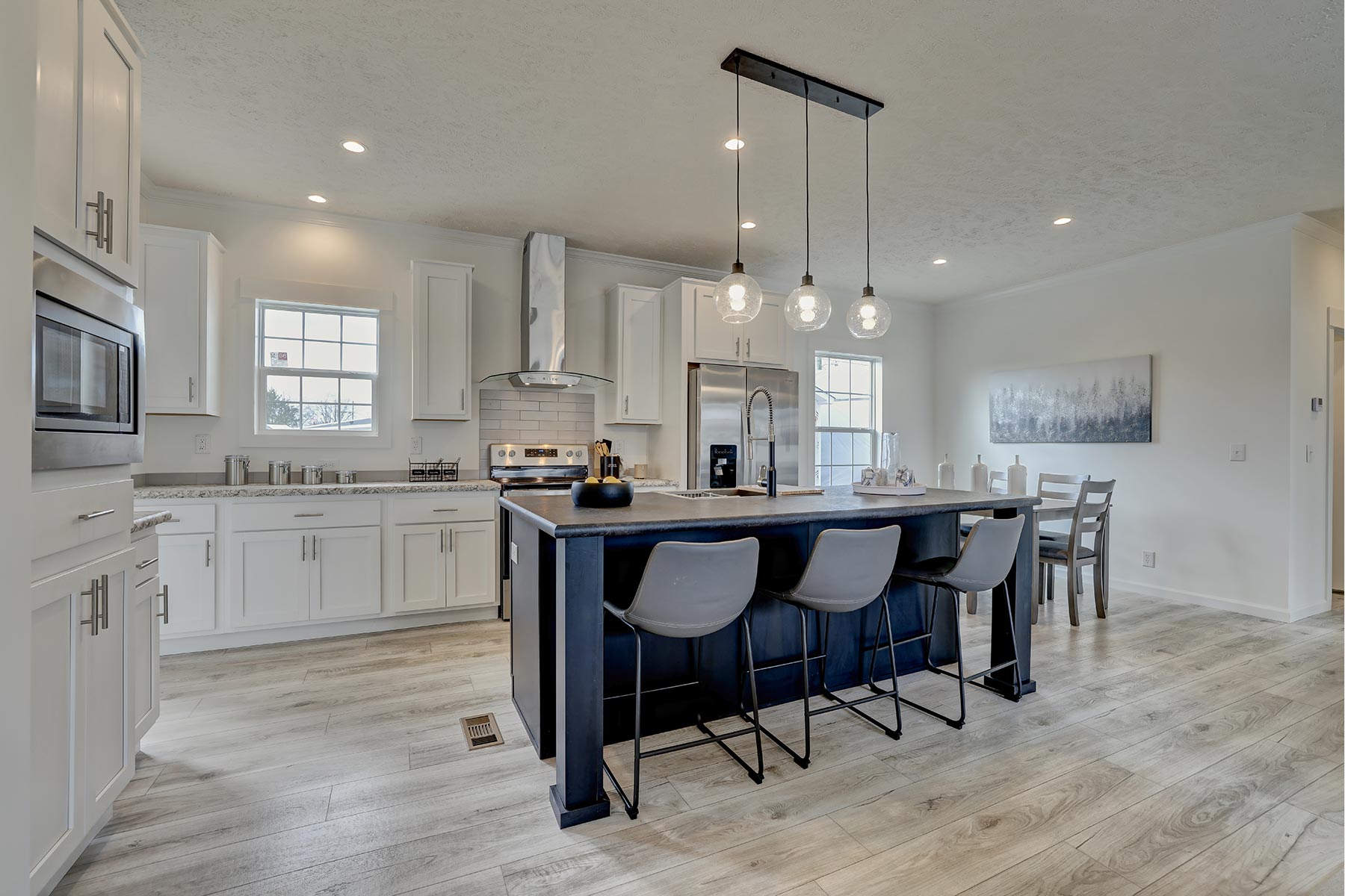 Modern kitchen with light wood floors, white cabinets, and a central island with gray chairs. Three pendant lights hang above, creating a cozy atmosphere.