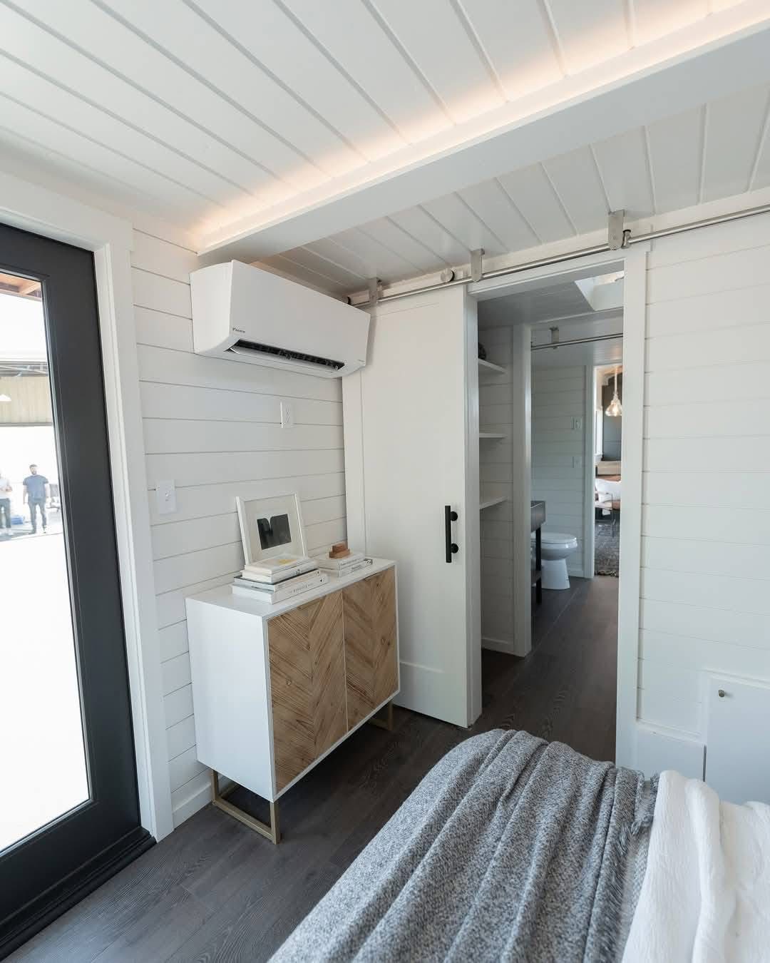 Compact modern bedroom with white panel walls, a sliding barn door revealing a bathroom, a small cabinet with decor, and a cozy bed, creating a serene atmosphere.