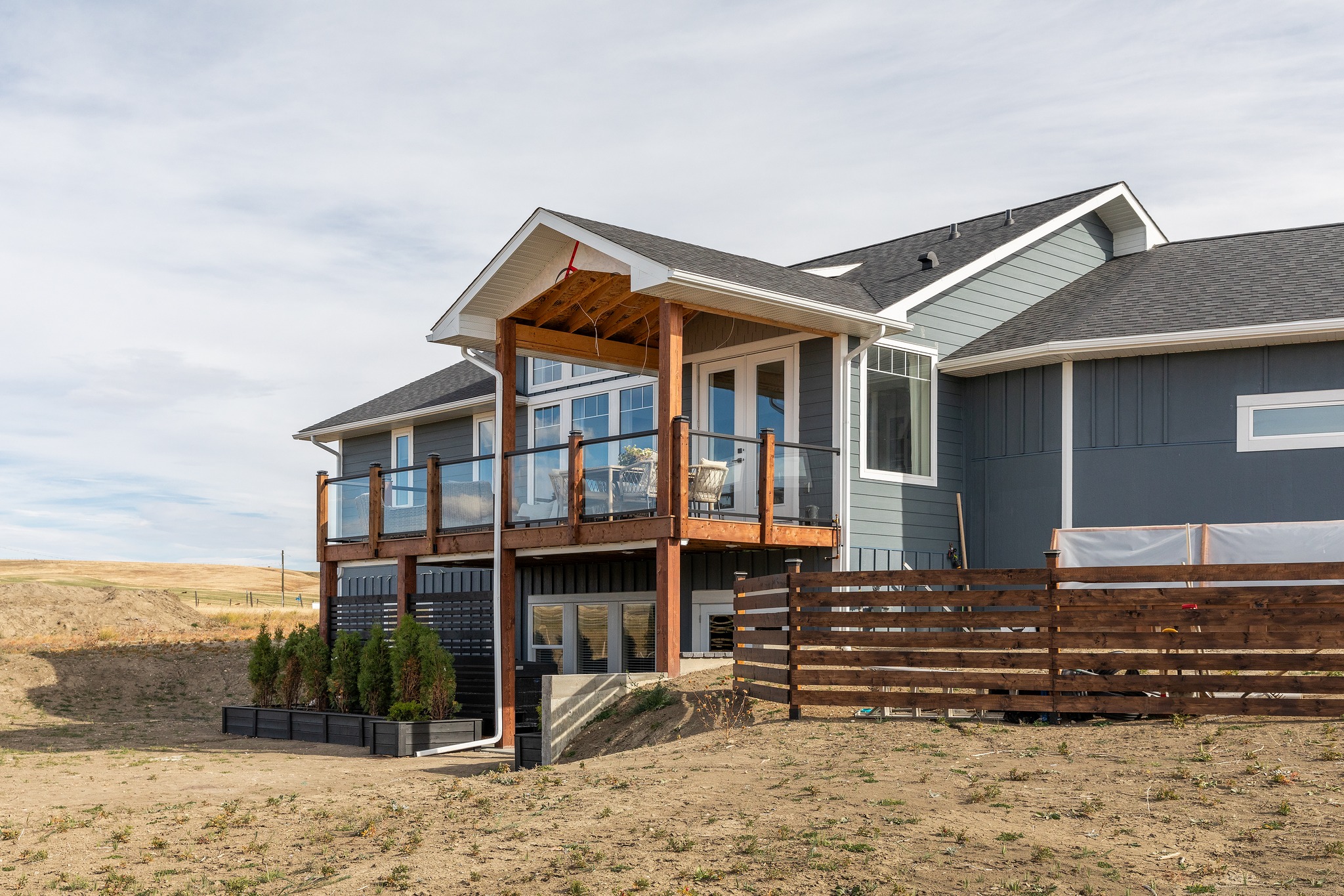 Modern two-story house with a wooden balcony, large windows, and a wooden fence. Set in a barren, open landscape under a cloudy sky.