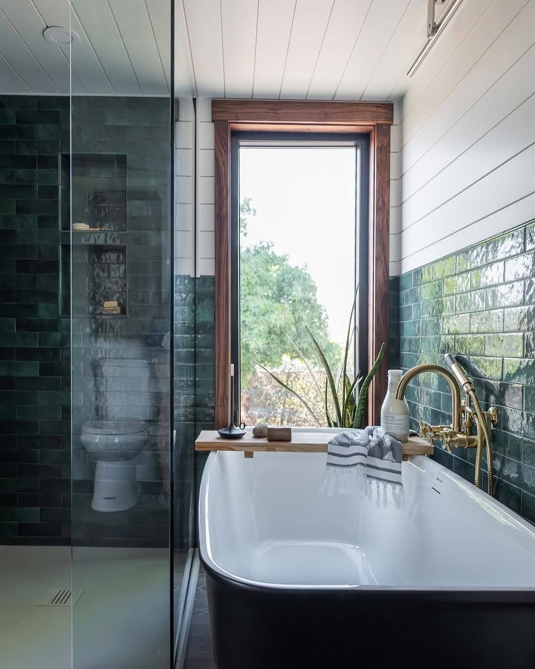 Modern bathroom with a white freestanding bathtub, brass fixtures, and green tile walls. A large window offers a view of trees, creating a serene atmosphere.