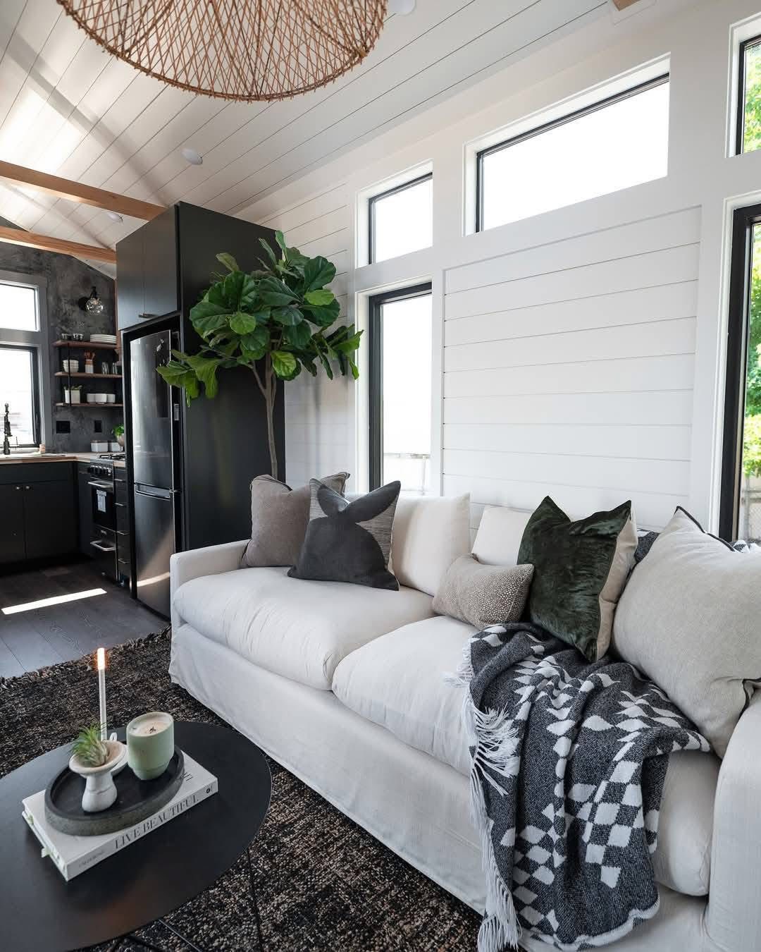 Cozy living room with a white sofa adorned with cushions and a patterned throw. A large plant and modern kitchen create a warm, inviting atmosphere.