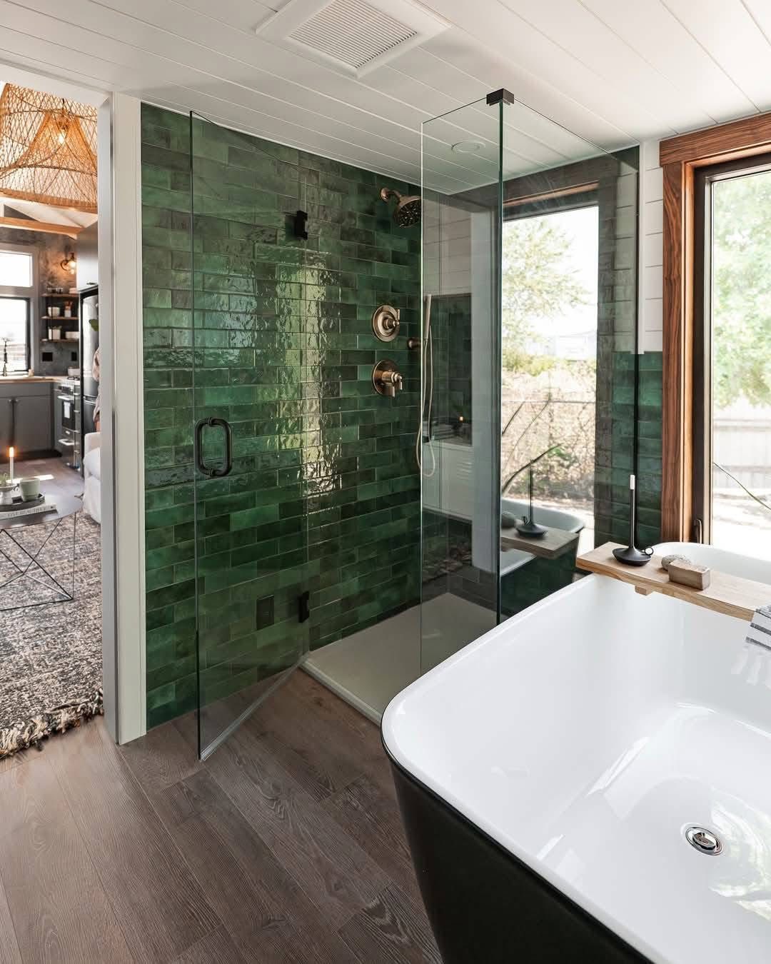 Modern bathroom with green-tiled shower, glass door, and brass fixtures. Freestanding white tub with wooden caddy, large window, bright and inviting.