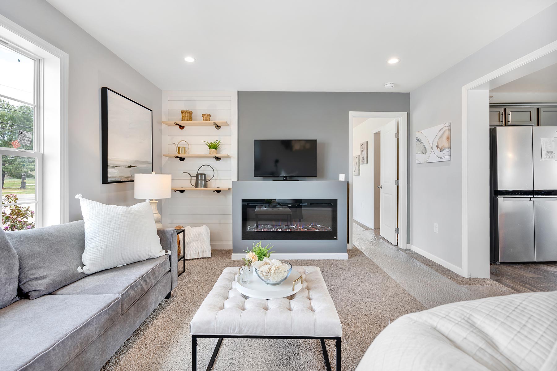 Modern living room with a gray sofa, white pillows, and a tufted ottoman. A wall-mounted TV and electric fireplace provide a cozy ambiance.