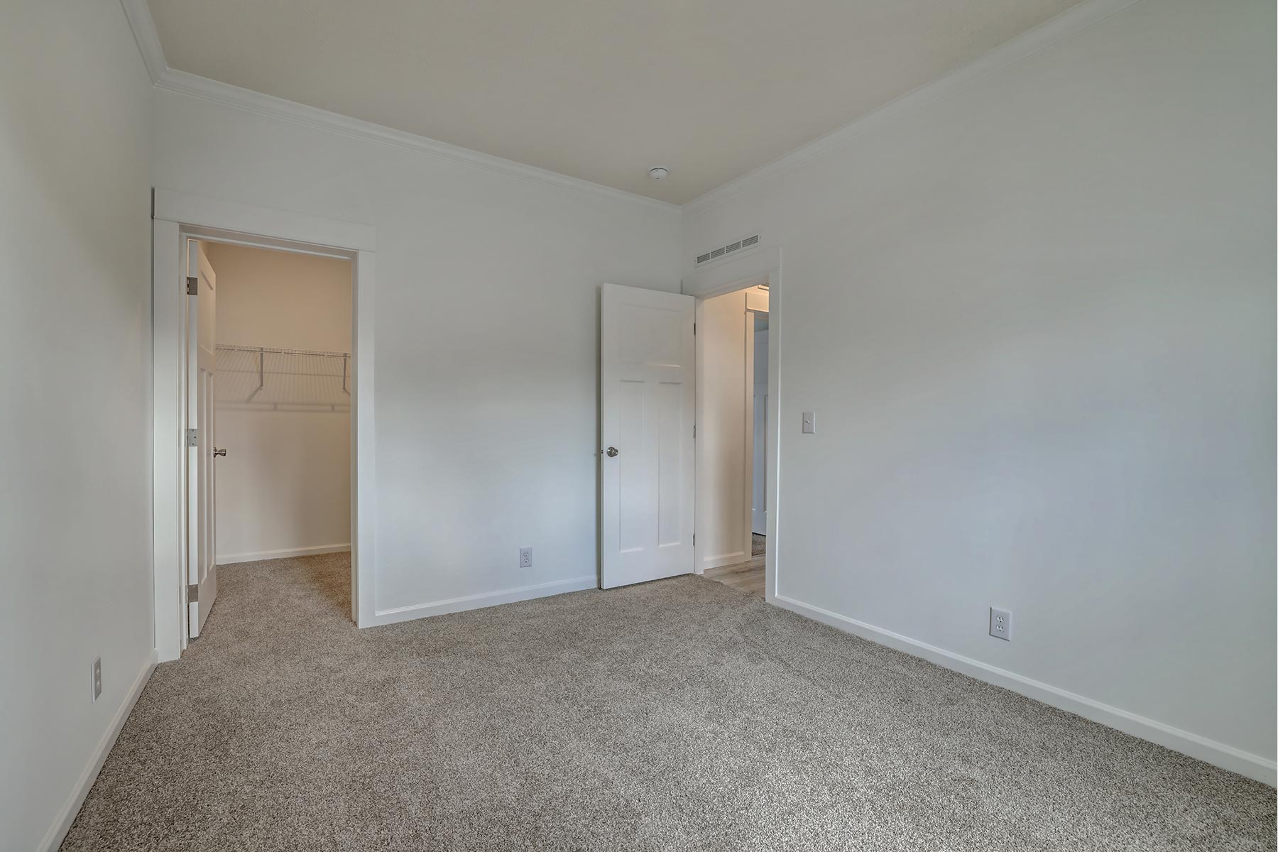 Empty room with light gray carpet and white walls. Two open doors reveal a closet and a hallway, creating an inviting and open atmosphere.