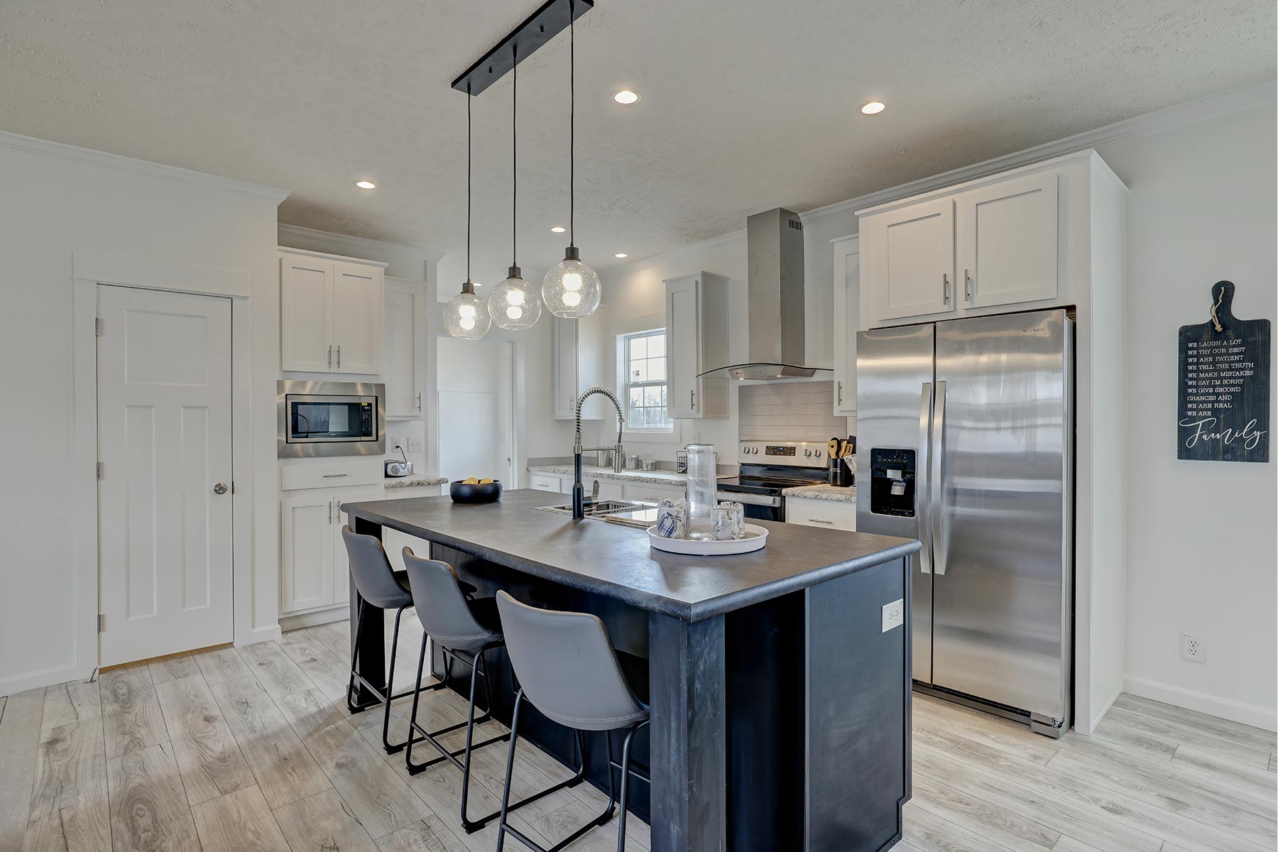 Modern kitchen with a black island, high stools, pendant lights, and stainless steel appliances. Light wood floors and white cabinets add brightness.